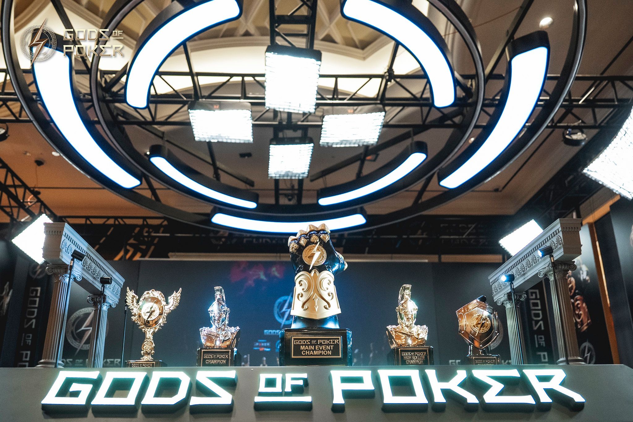 Multiple poker trophies on display under a bright circular light, with 'GODS OF POKER' text.