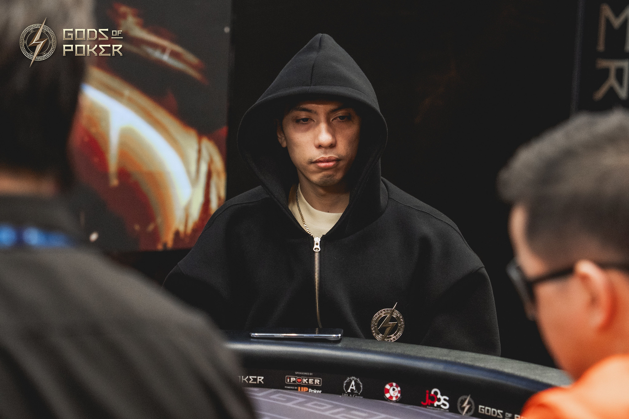 A serious man in a black hoodie sits intently at a poker table during a game.