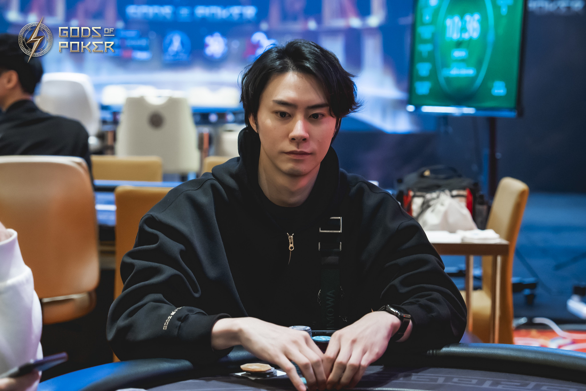 A focused young man in a black hoodie sits at a poker table with chips.
