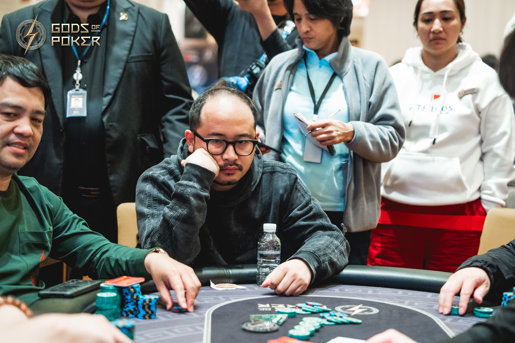 A man in glasses thoughtfully considers his poker hand at a busy casino table.