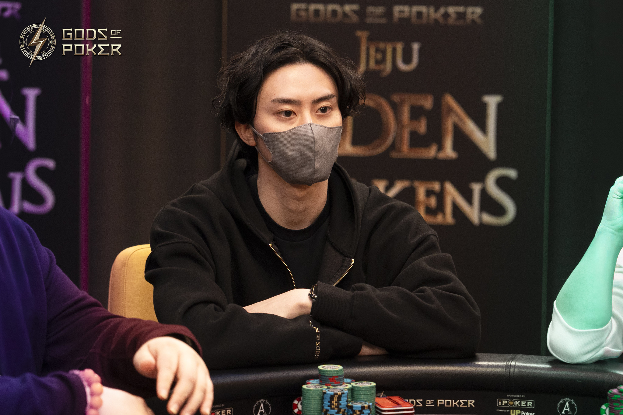 Man in a black hoodie and face mask sits at a poker table, looking focused.