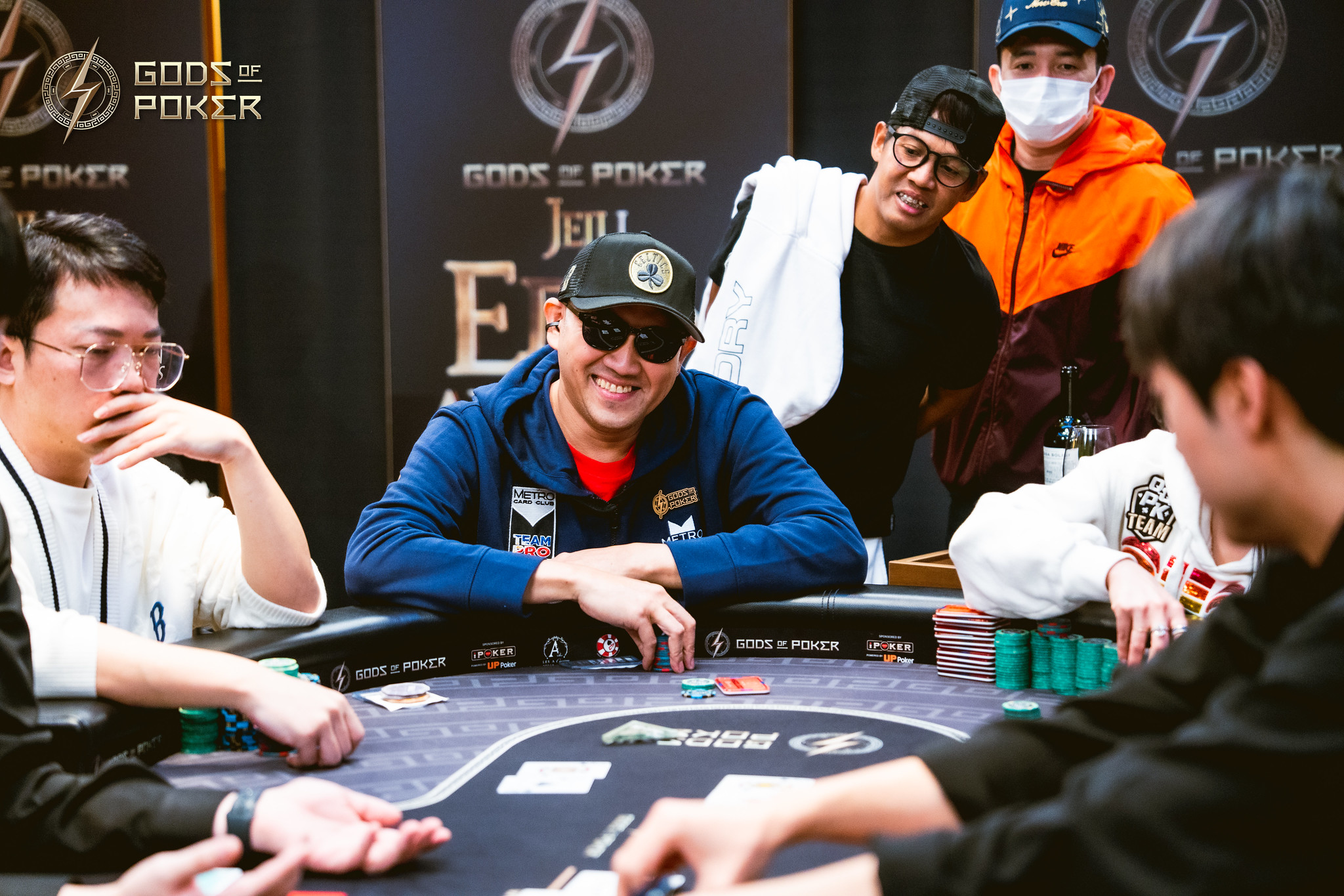 A man in sunglasses smiles cheerfully at a poker table surrounded by players and onlookers.