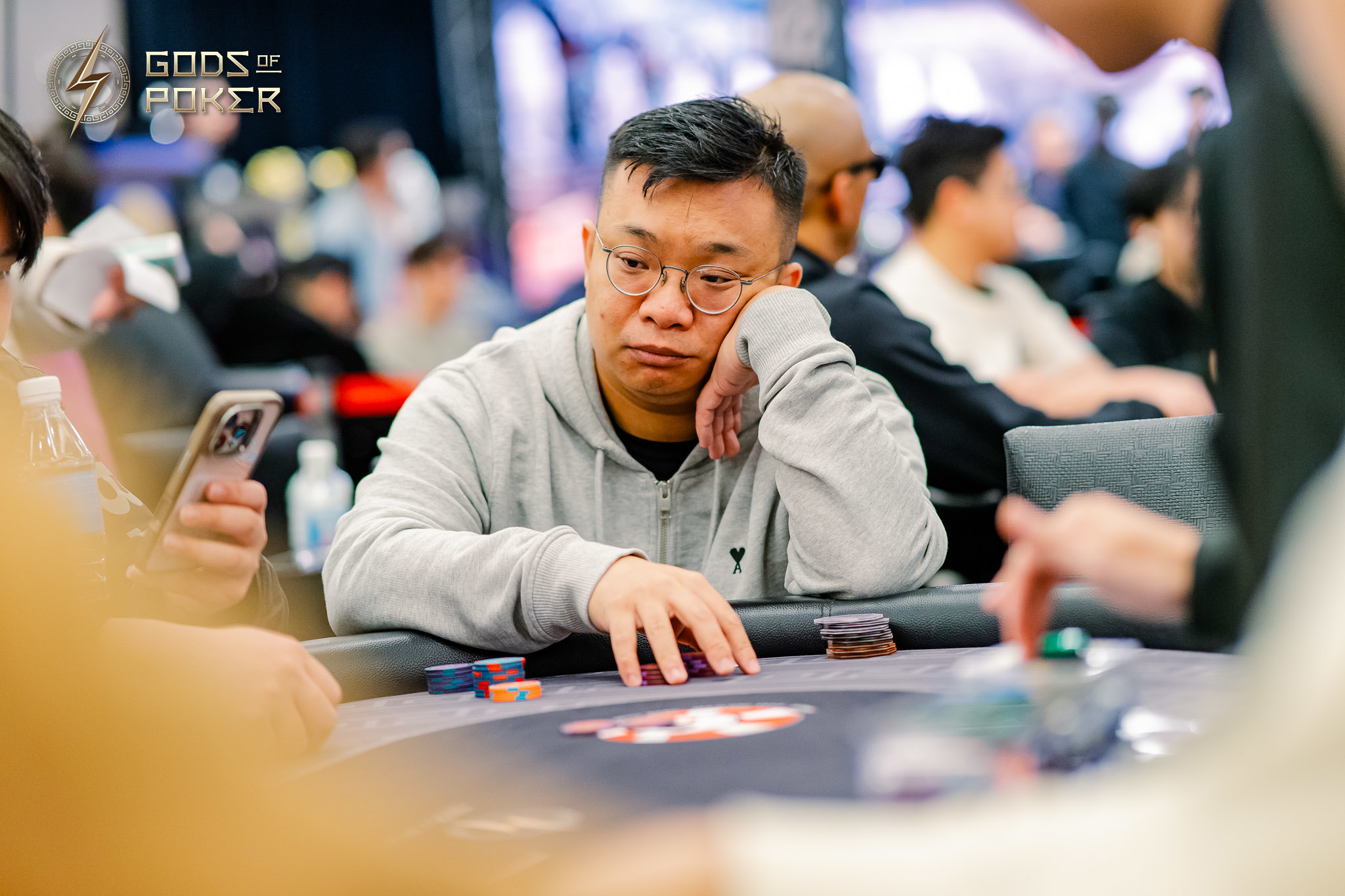 Man in grey hoodie sits at poker table, looking thoughtful with hand on his face.
