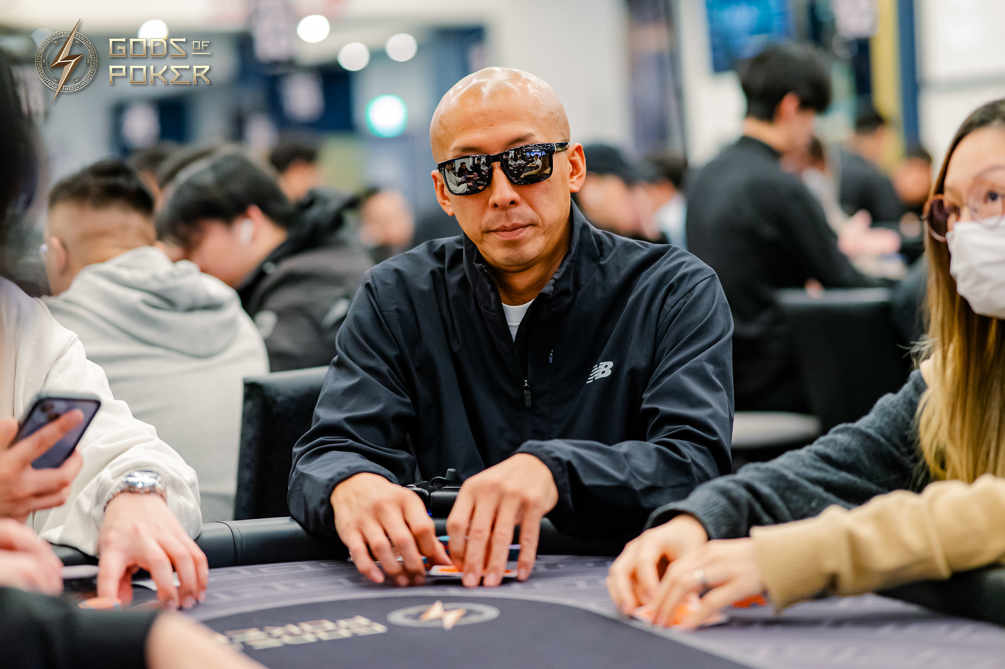 Bald man in sunglasses holding poker chips at a busy tournament table.