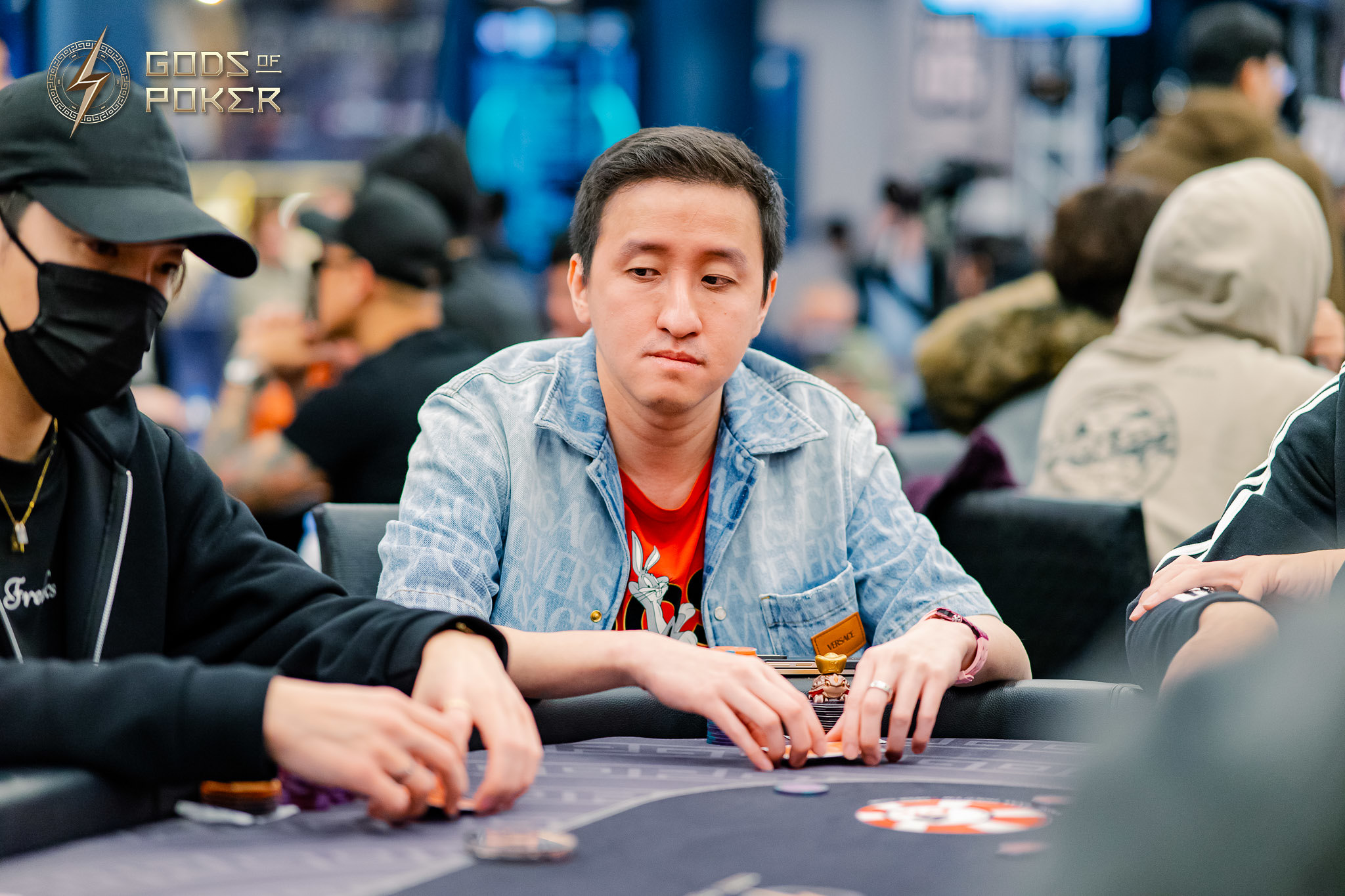 Man in denim jacket contemplating his hand at a busy poker table with chips.