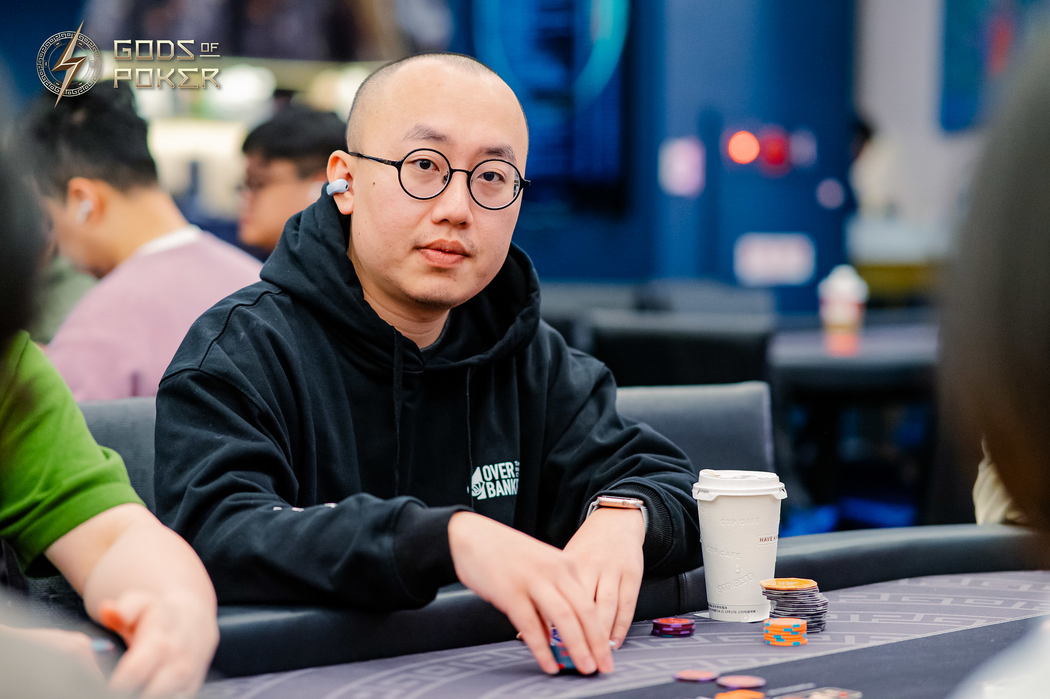 Bald man with glasses and black hoodie playing poker, chips on table.