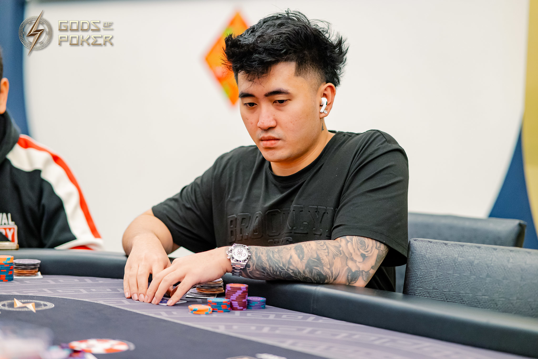 Young man with tattoos and earbuds playing poker, focused on chips at the table.