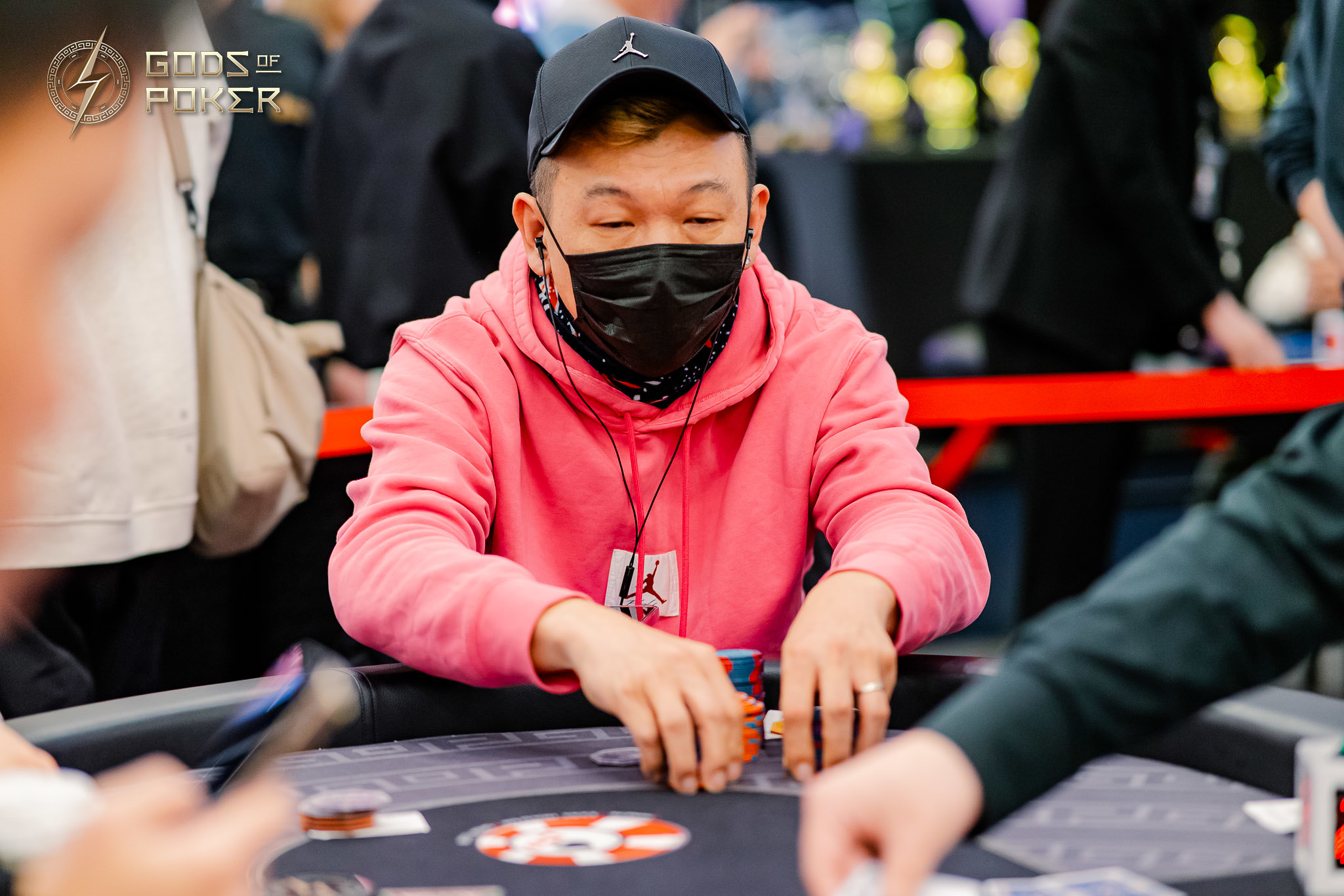Man in a pink hoodie and black mask playing poker, moving chips on the table.