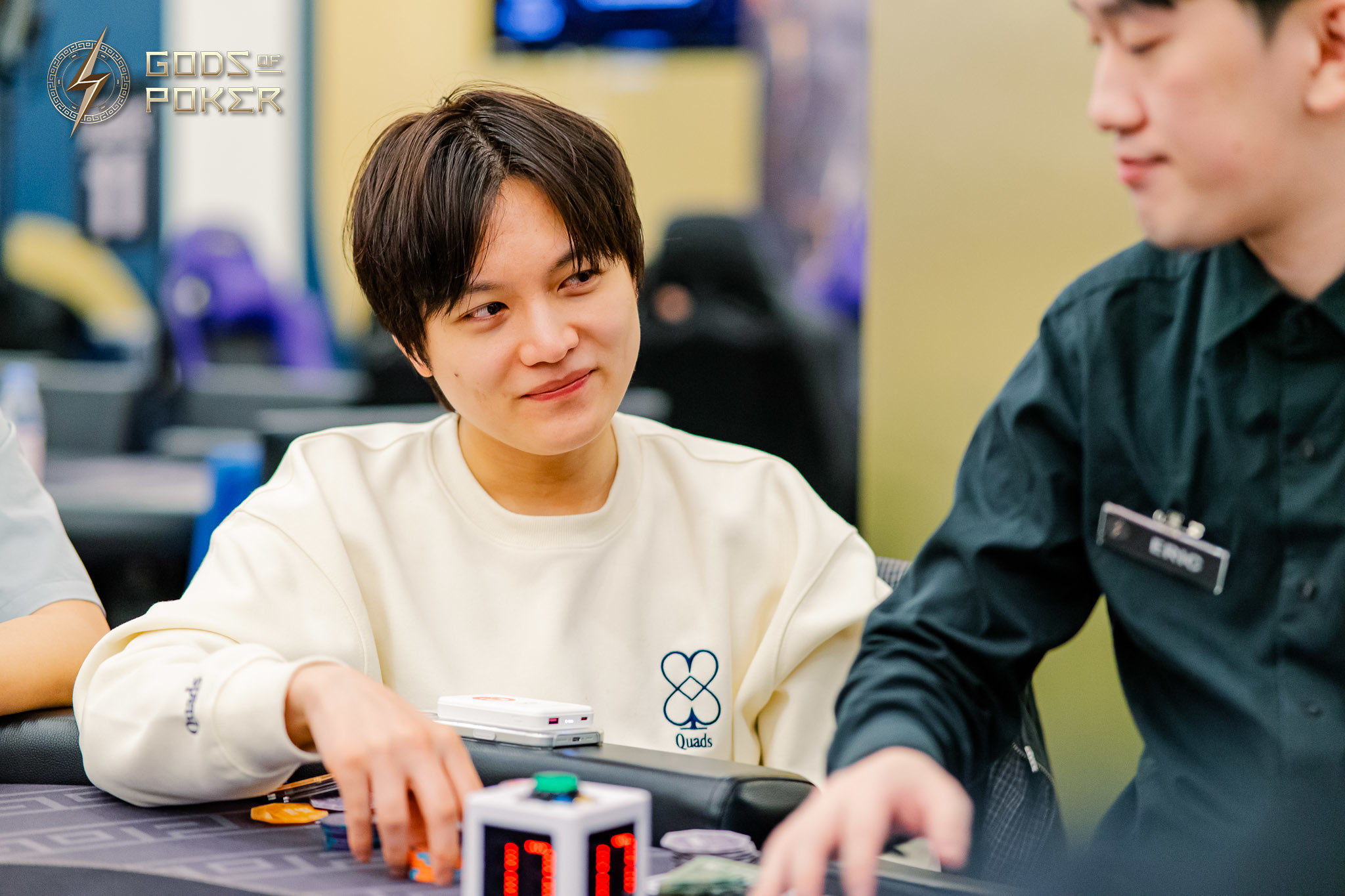 A person in a light sweatshirt smiles while seated at a poker table with chips.