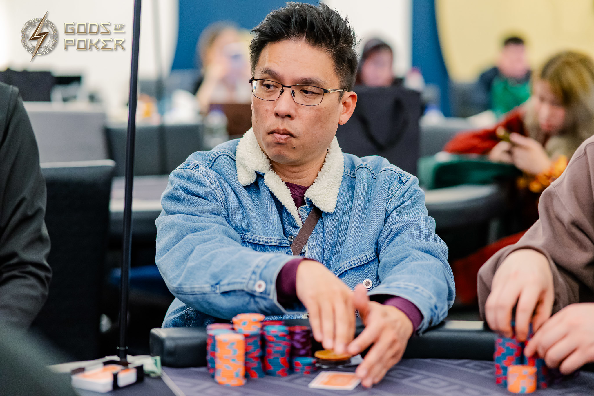 A man wearing glasses and a denim jacket concentrates at a poker table with chips.