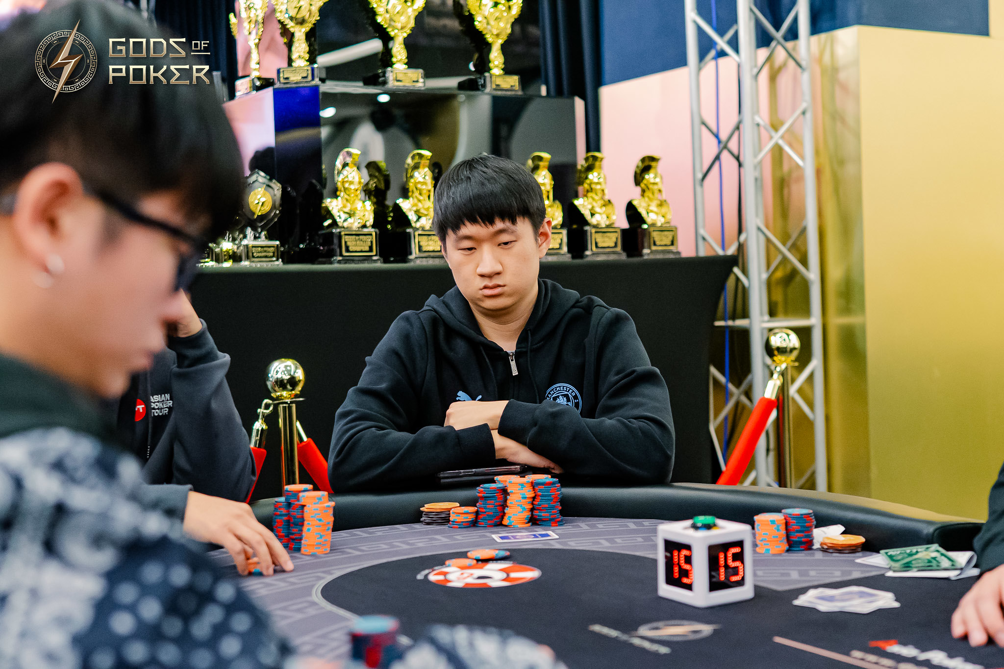 An Asian man in a black hoodie sits focused at a poker table surrounded by chips.