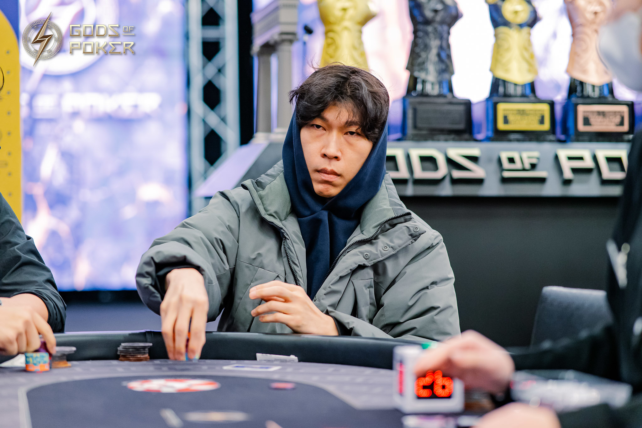 A man in a hooded jacket intently plays poker, moving chips on a dark table.