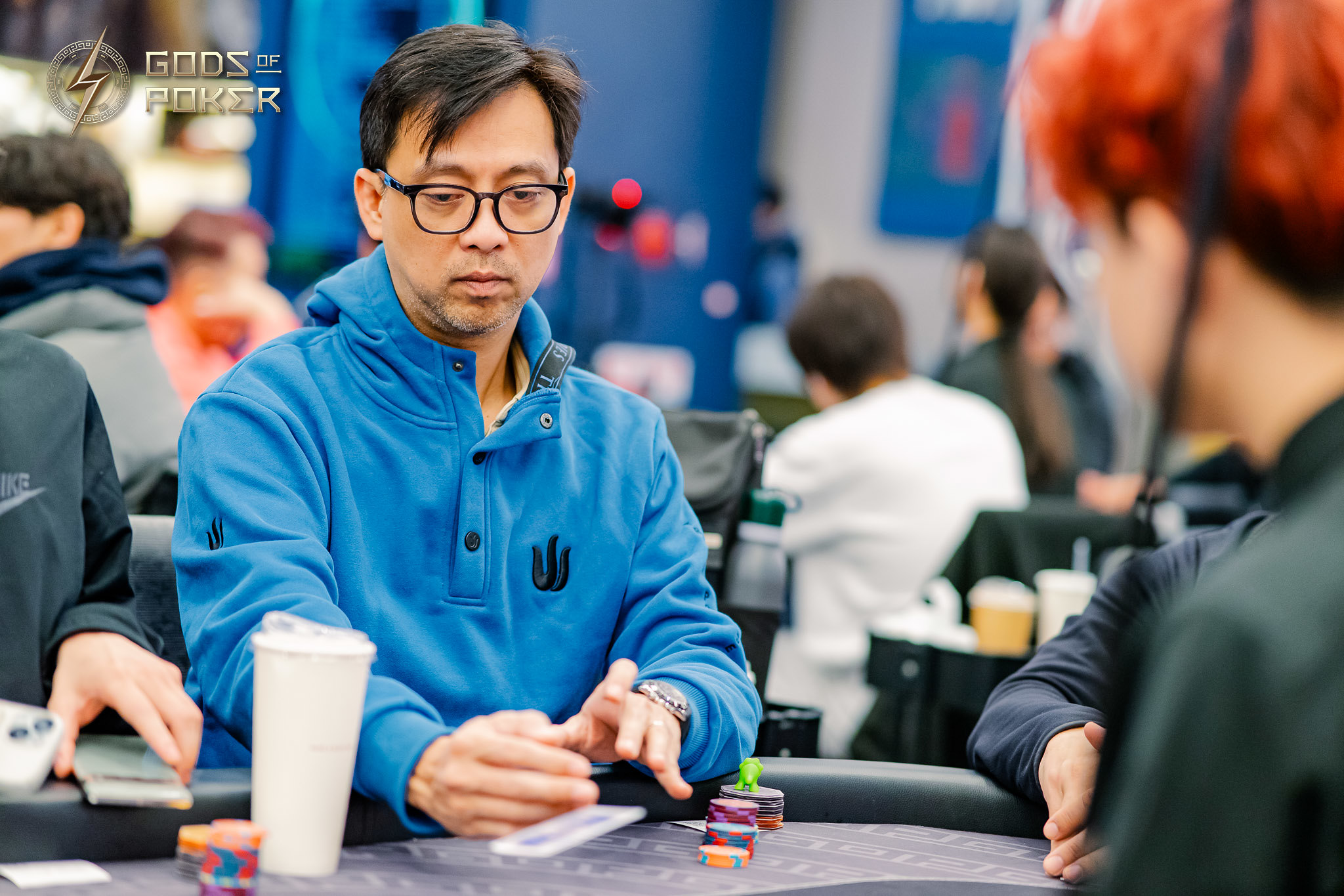 Man in glasses and blue hoodie intently playing poker, looking at his cards.
