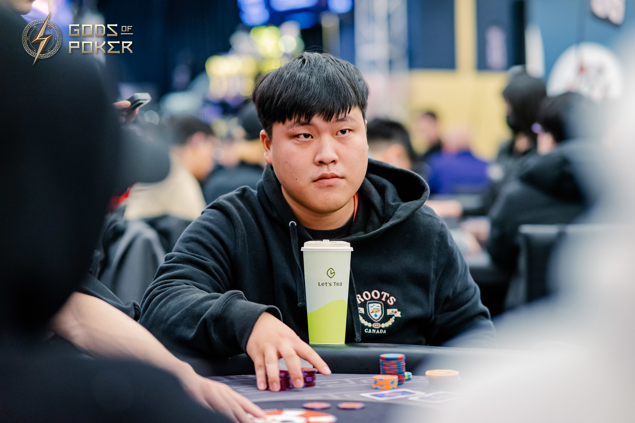 Young man in black hoodie at poker table with serious expression, chips, and a drink.