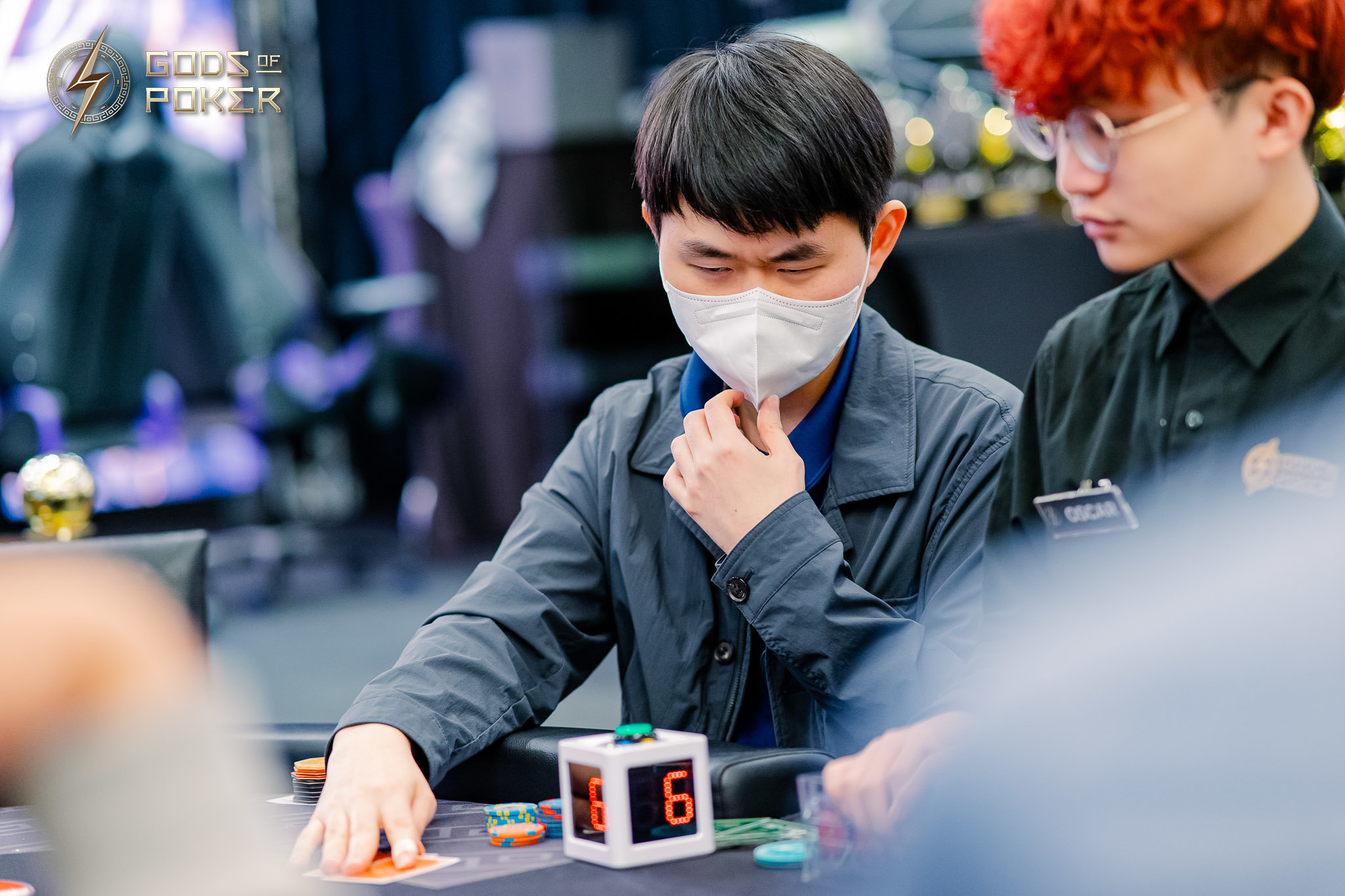 Man in face mask playing poker, looking at cards with a timer cube on table.
