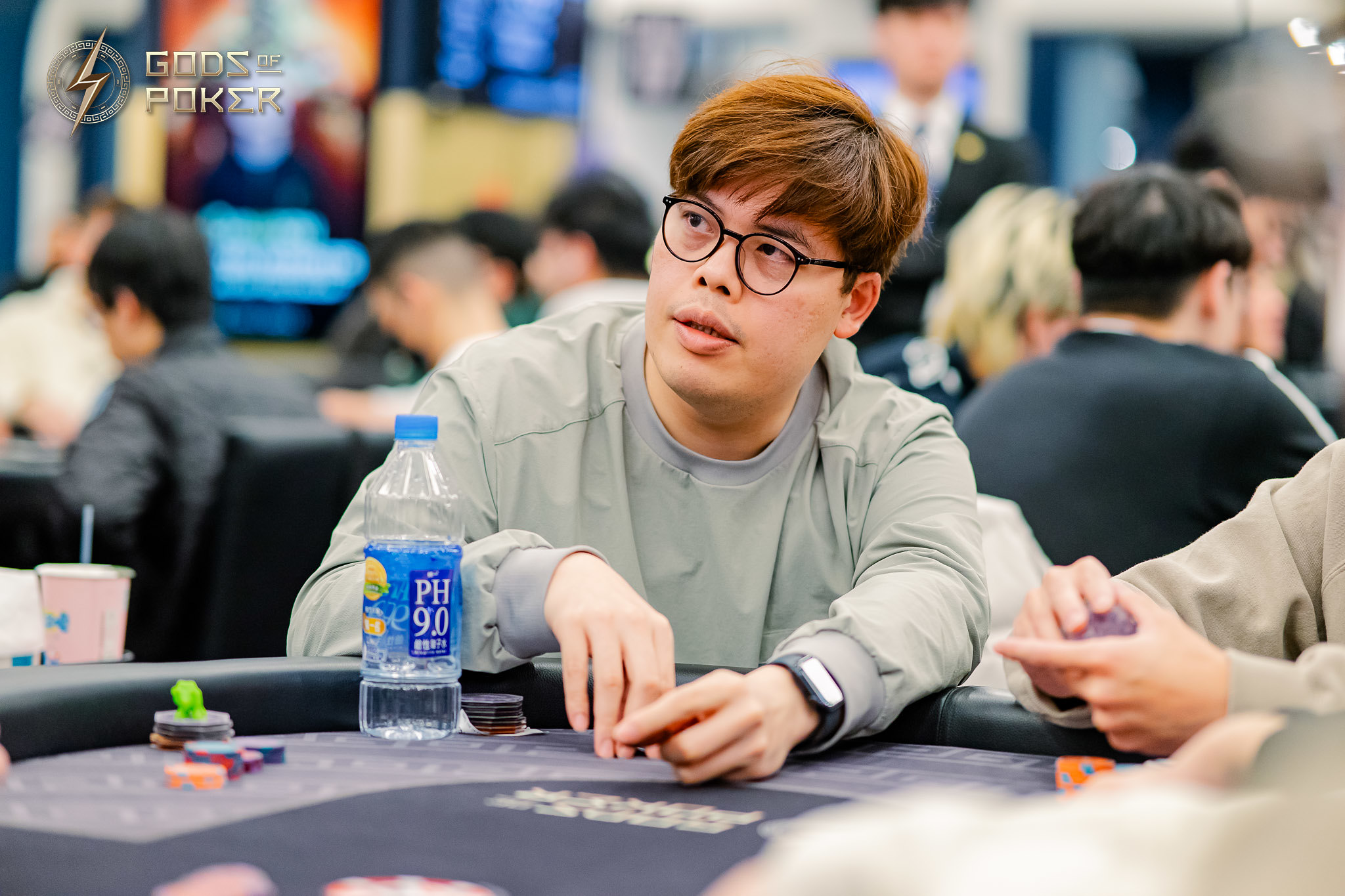 A man in glasses intently watches a poker game, holding his cards.