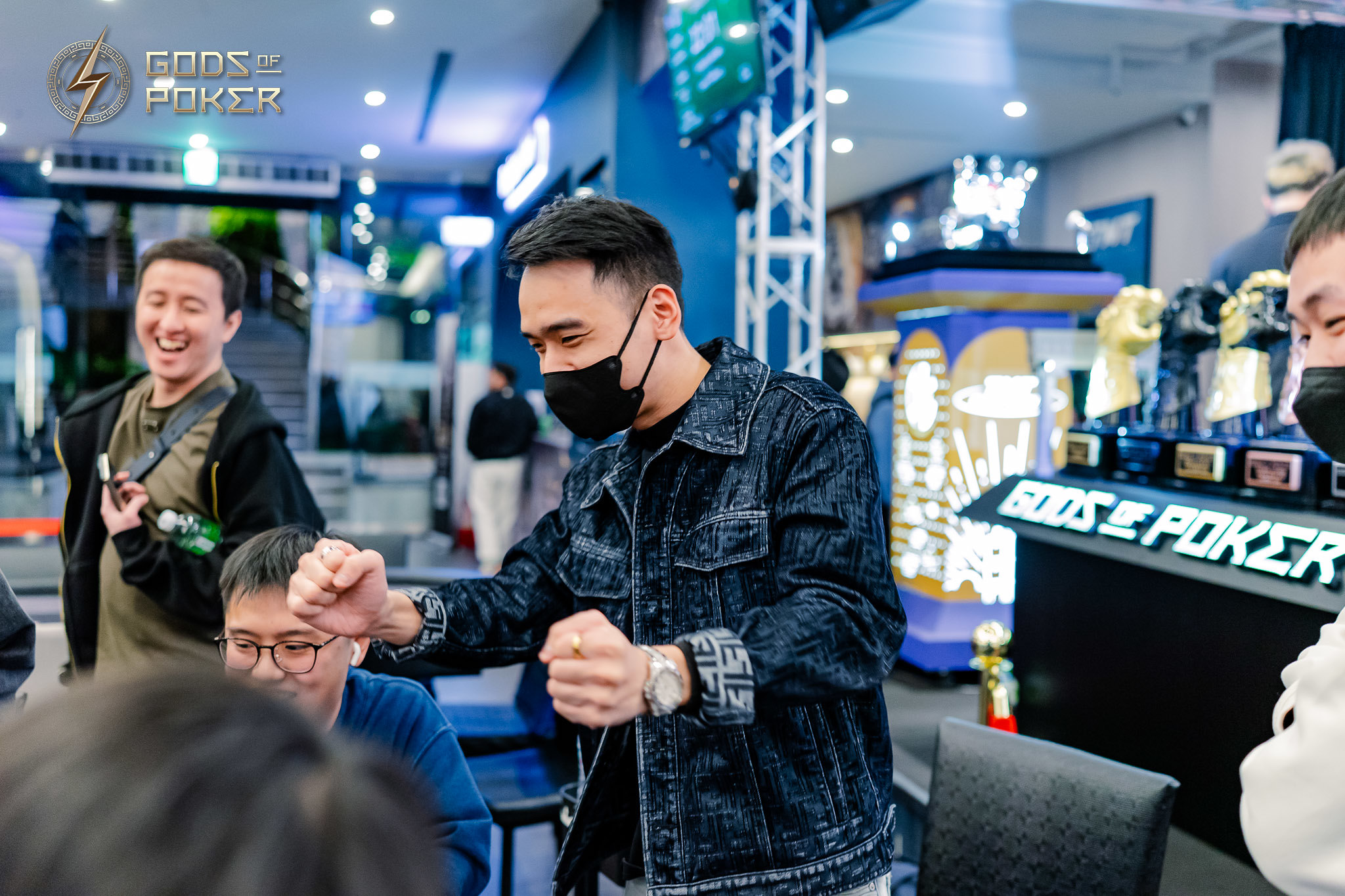 Man in denim jacket and face mask gestures excitedly at a poker table during a game.