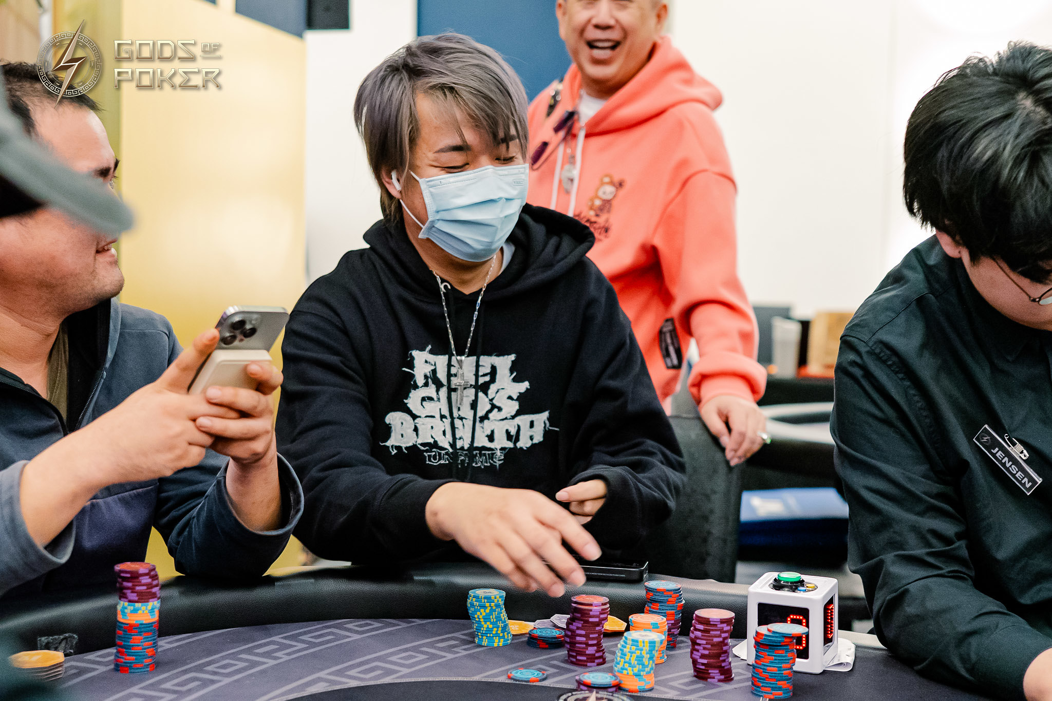 Masked player with gray hair moves poker chips at a lively card table.