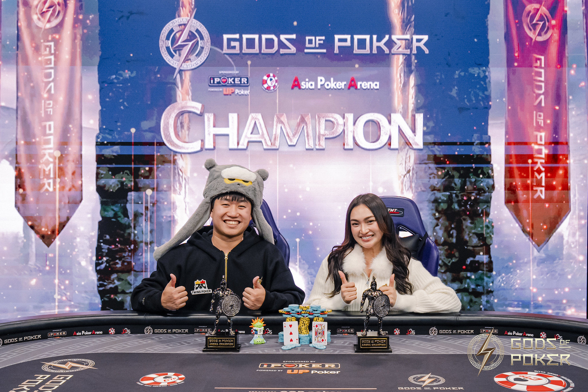 Two poker champions, a man in a fun hat and a woman, smiling proudly with their trophies.