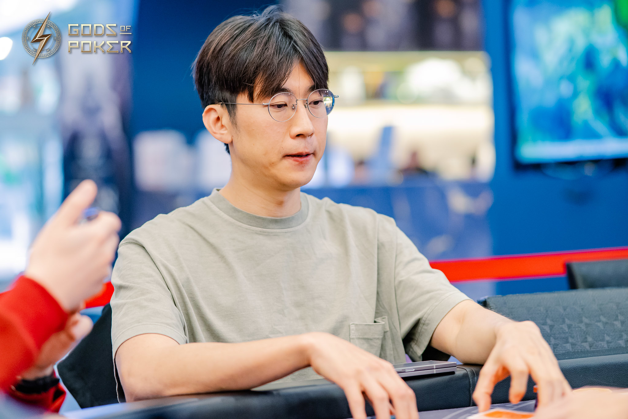 Asian man in glasses and a t-shirt sitting at a poker table, intently focused.