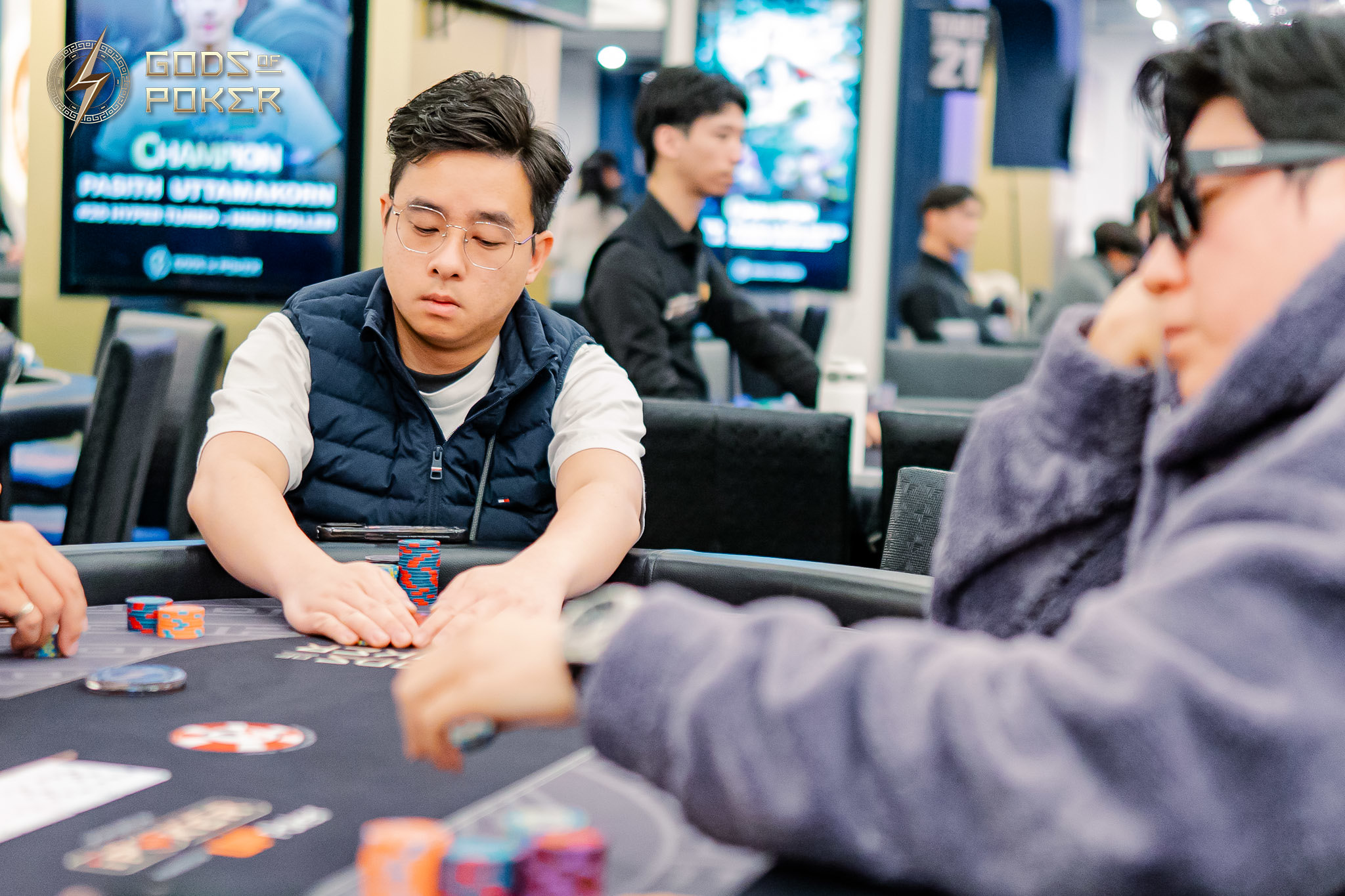 Man in blue vest playing poker, intently looking at his cards at the table.