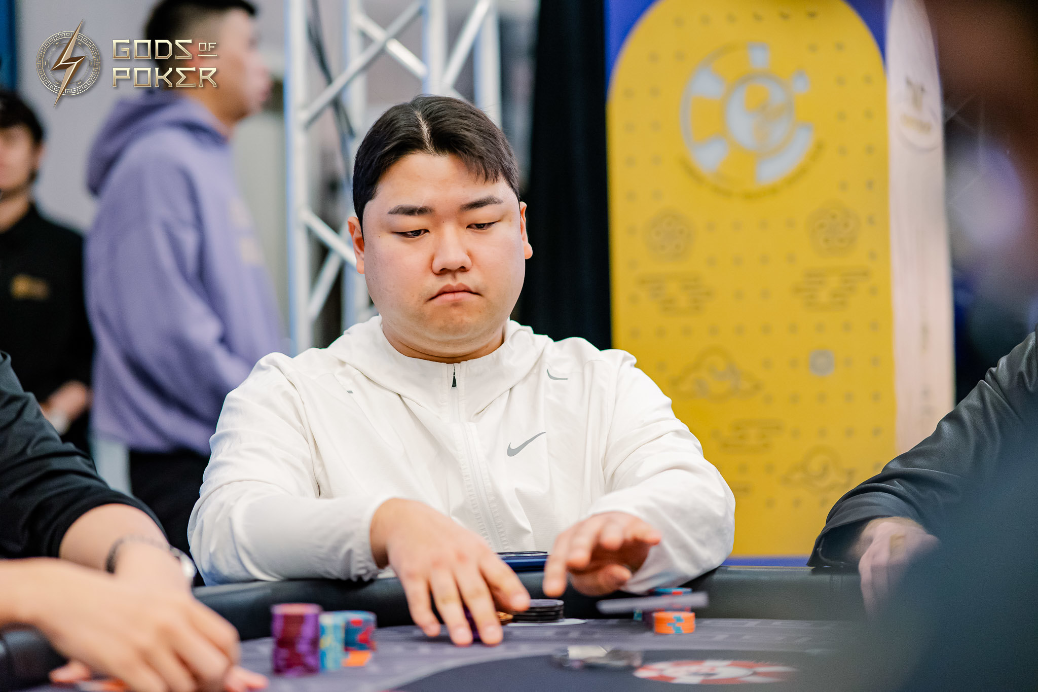 East Asian man in white jacket intently plays poker at a table with chips.