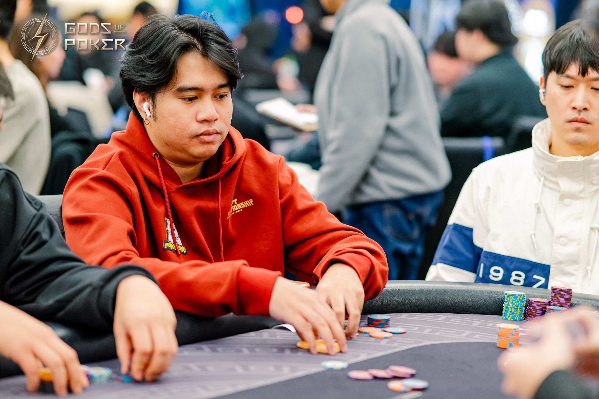 Man in red hoodie playing poker, moving chips at a table with other players.