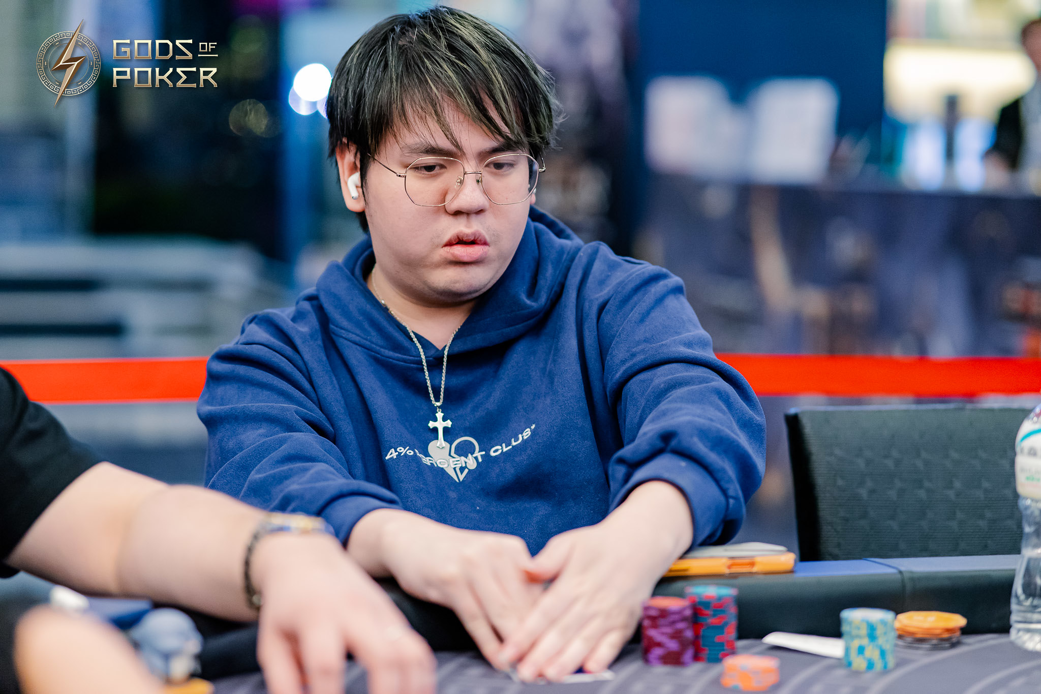 Man in blue hoodie with glasses intently watches poker game, hands on chips.