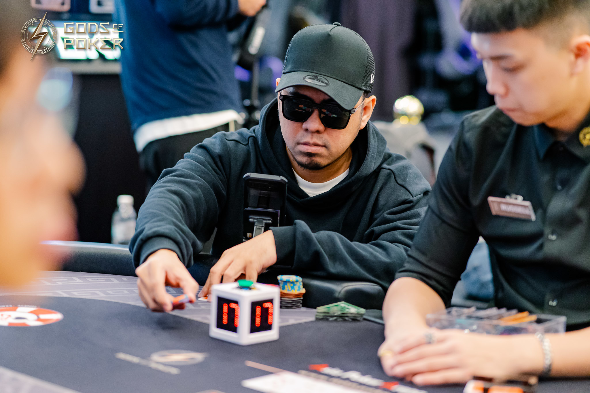 Man in sunglasses and black hoodie making a move during a poker game with a cube timer.