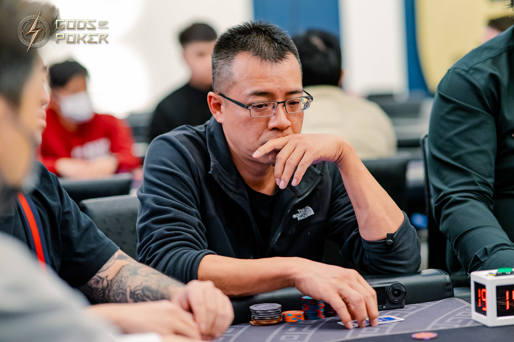 A man wearing glasses contemplates his hand at a poker table with chips.
