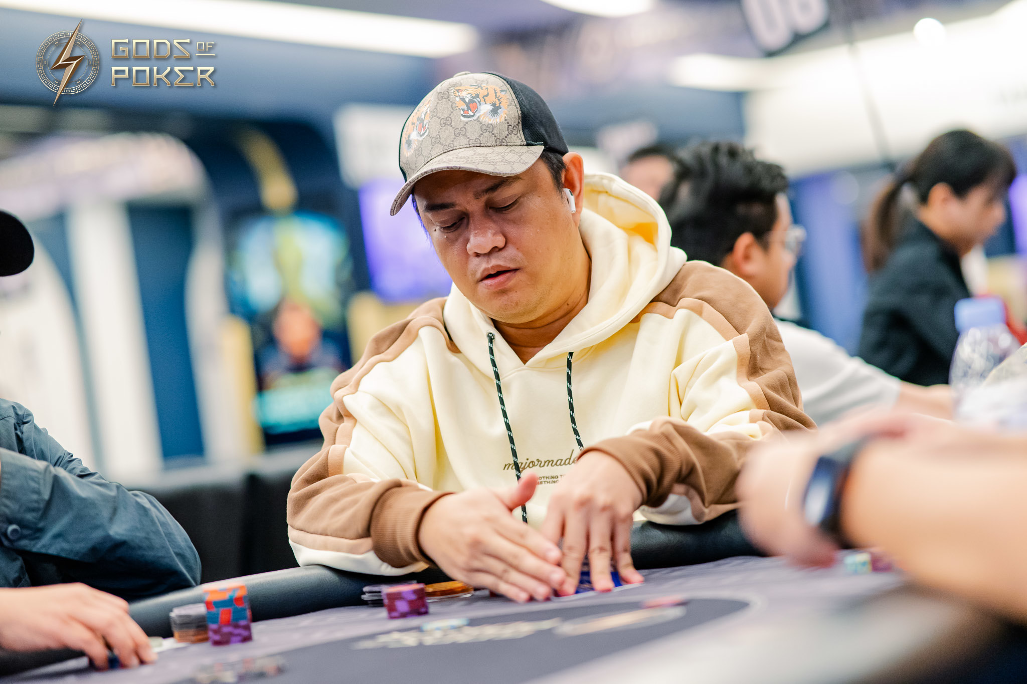 A man in a baseball cap and hoodie intensely plays poker, moving chips on the table.