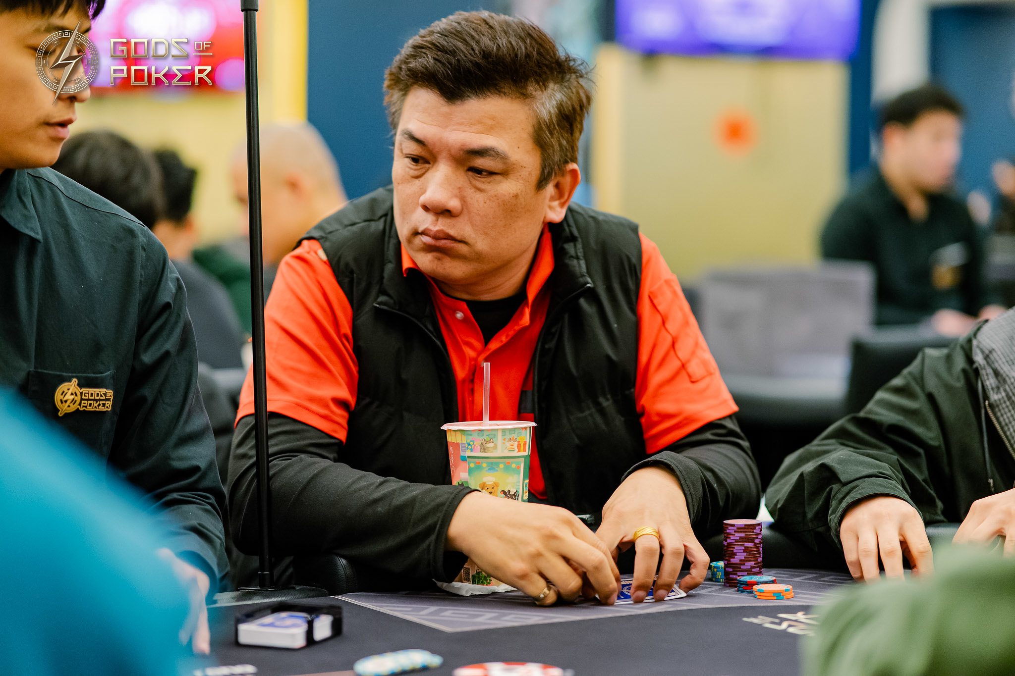 Man in black vest and red shirt intently playing poker at a table.