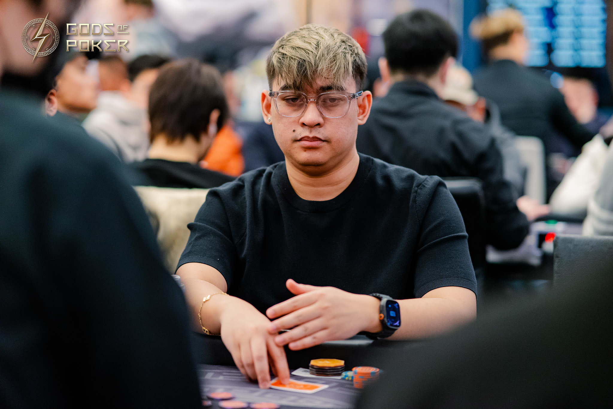 Young man in glasses and black shirt plays poker, focused on his hand.