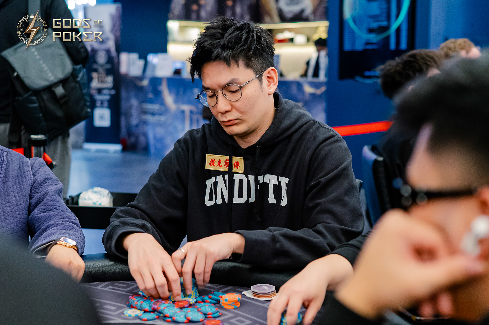 Man in black hoodie and glasses intently plays poker, looking down at his chips.