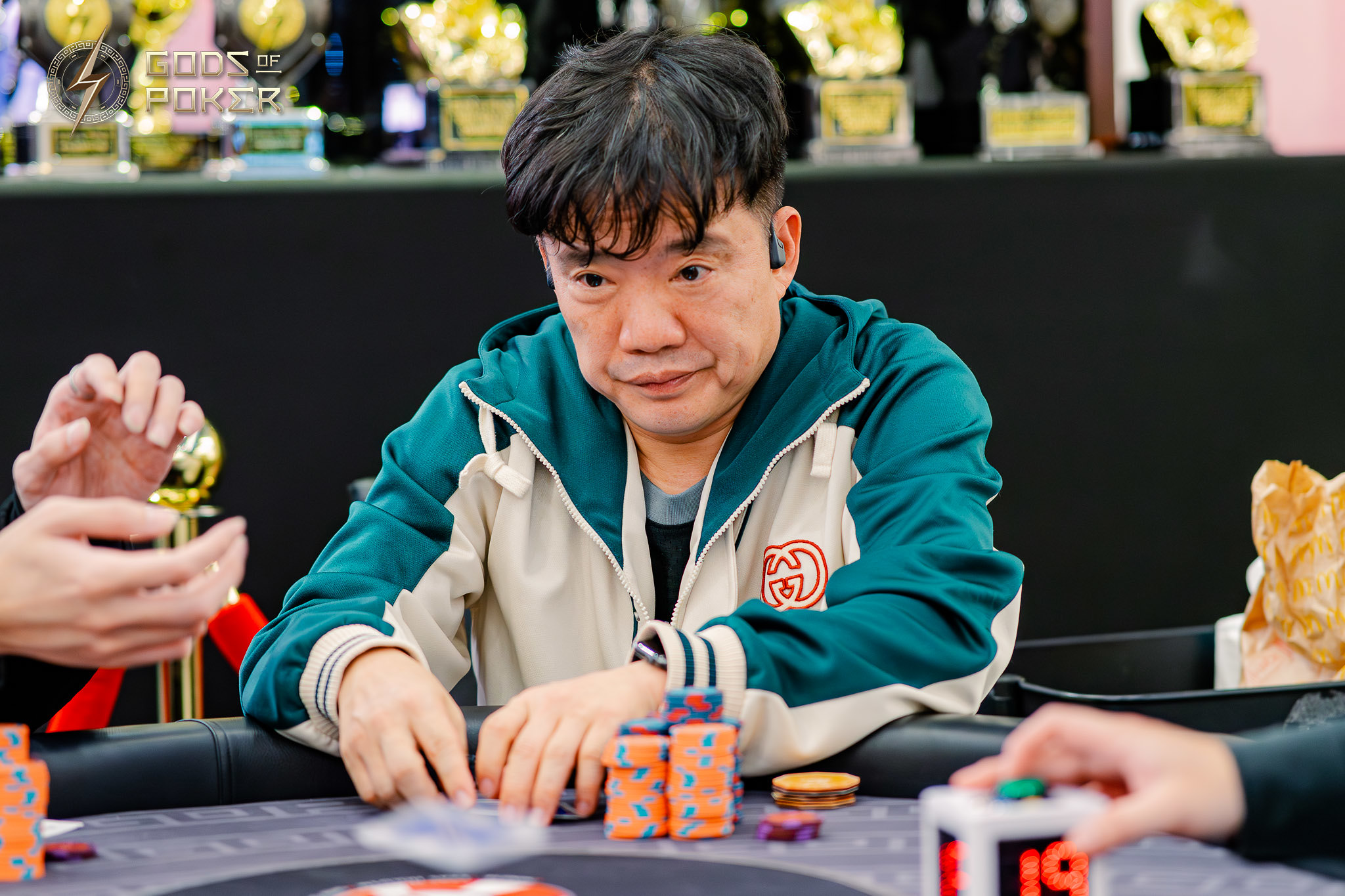 Asian man intently playing poker at a table with chips, trophies in the background.