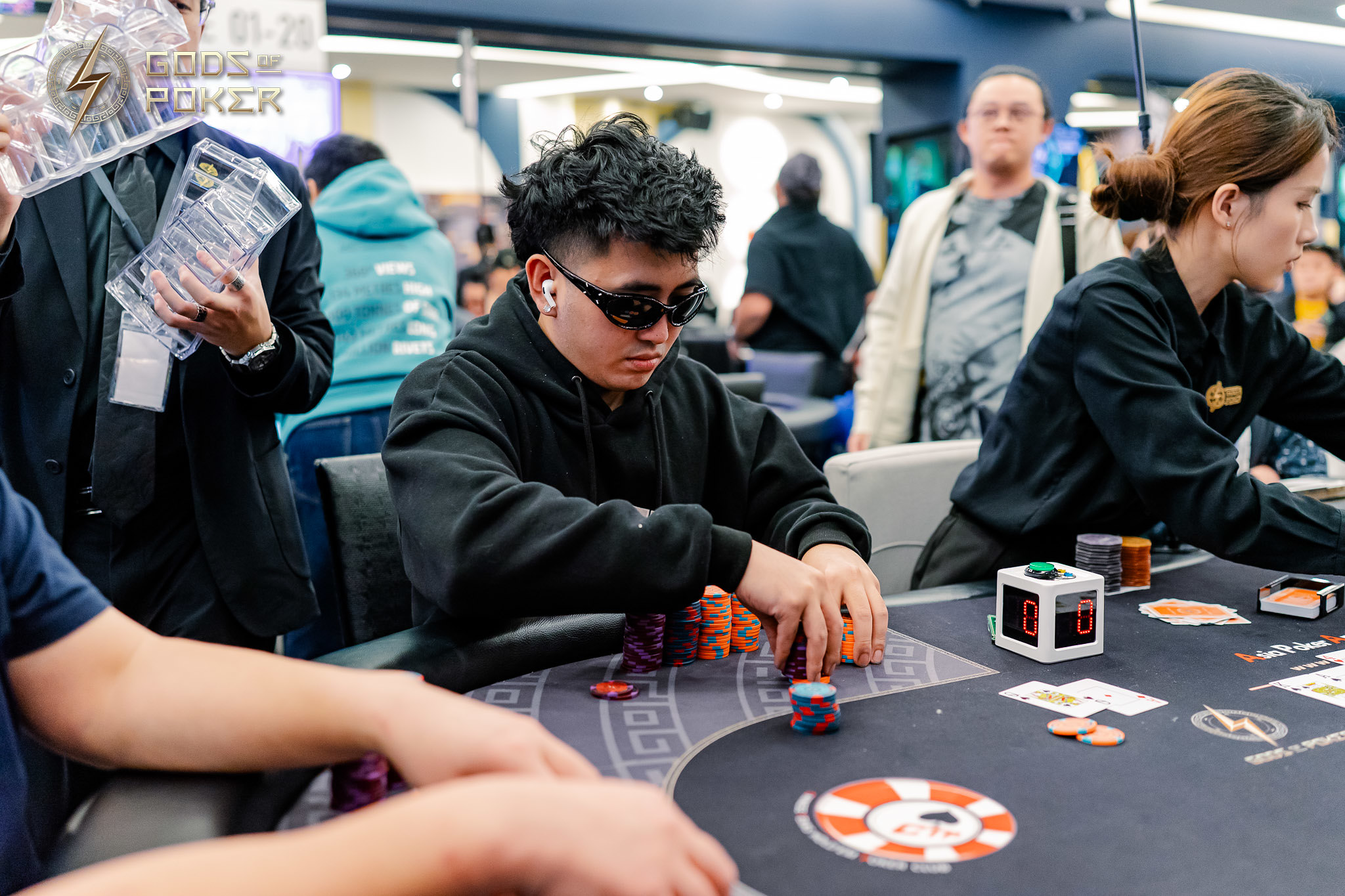 Young man in sunglasses plays poker at a casino table, surrounded by players.