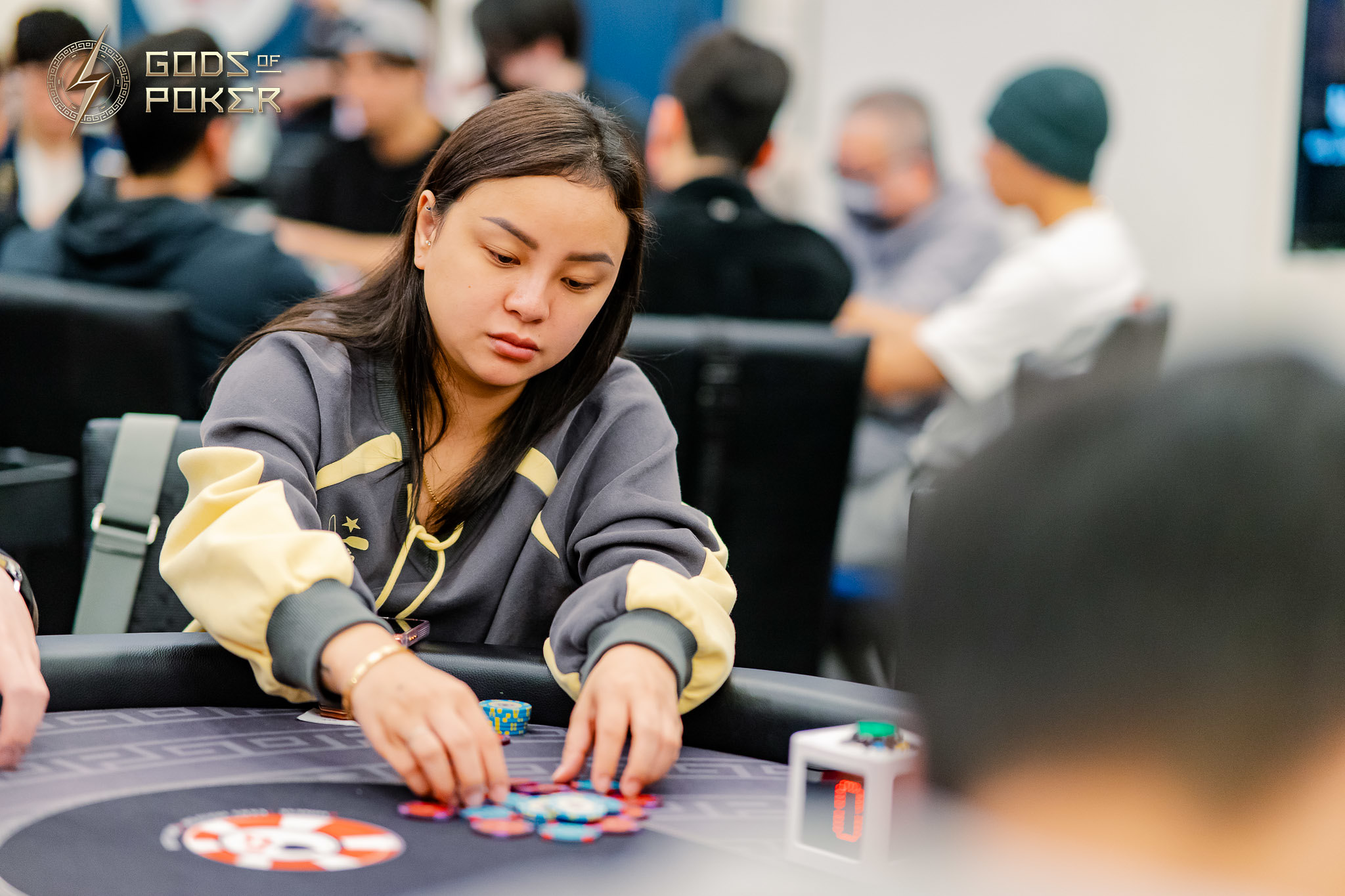A focused woman in a grey and yellow jacket plays poker, arranging chips on a green table.