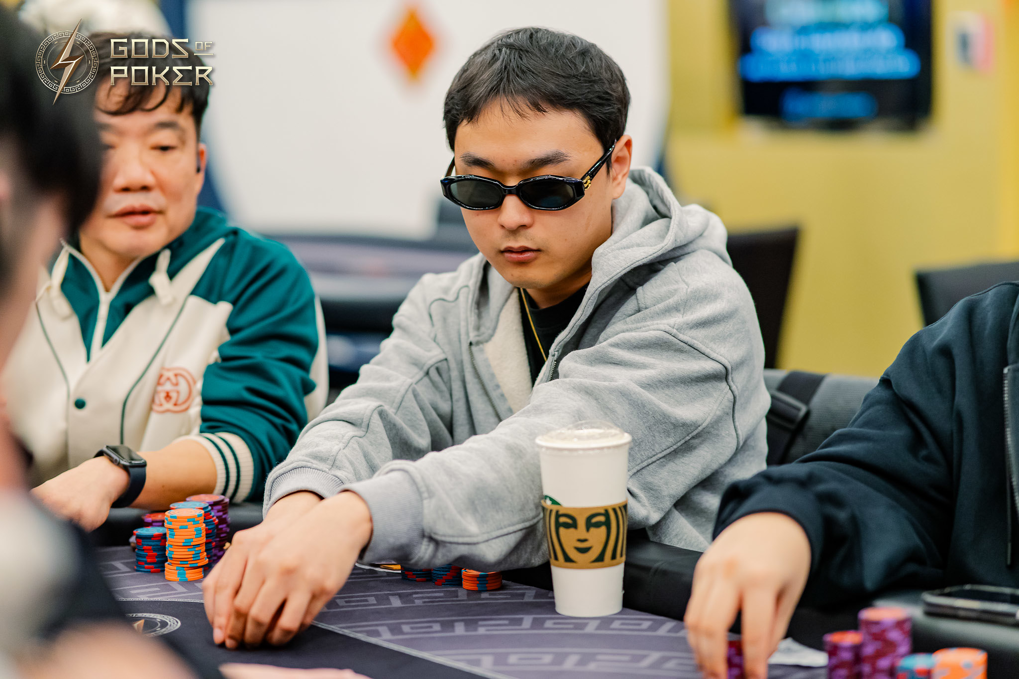 A man in sunglasses and a grey hoodie playing poker, moving chips on the table.