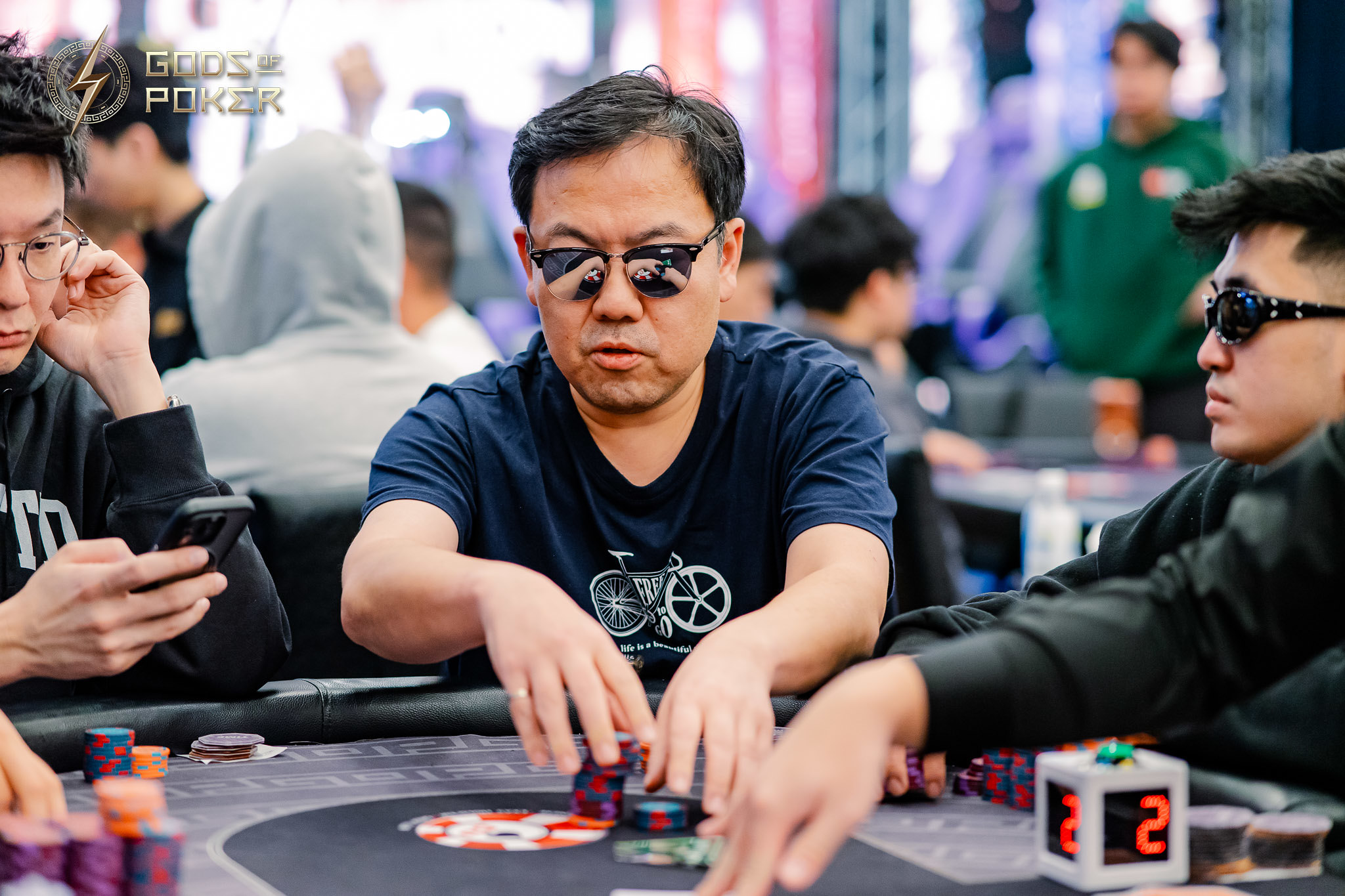East Asian man in sunglasses playing poker, moving chips at a casino table.