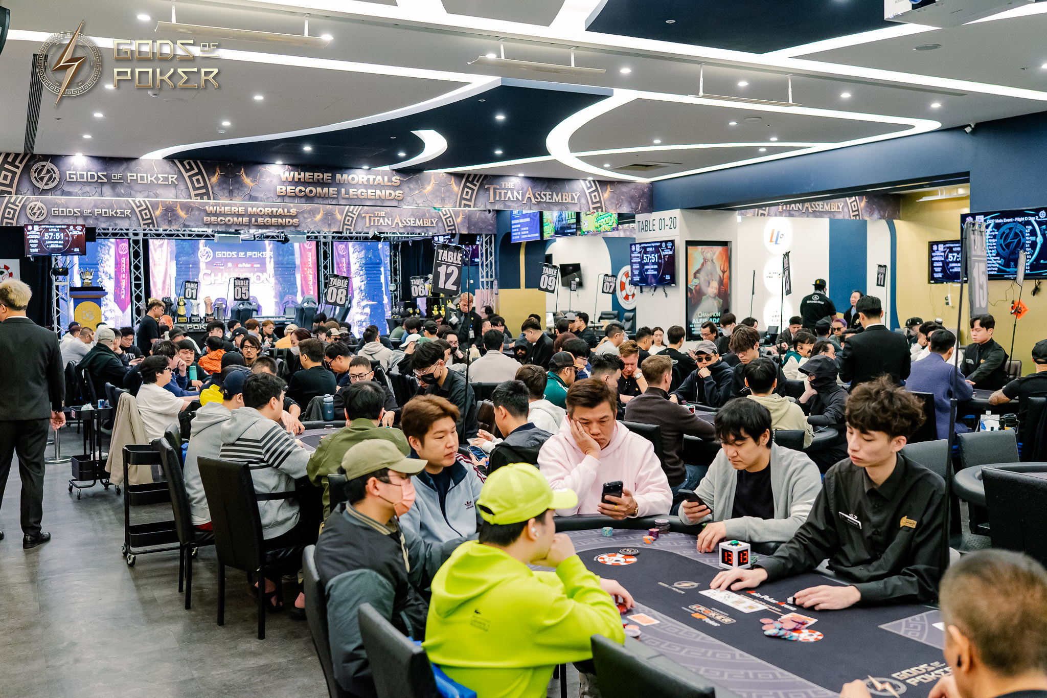 Many people are actively playing poker at tables in a crowded, modern casino room.