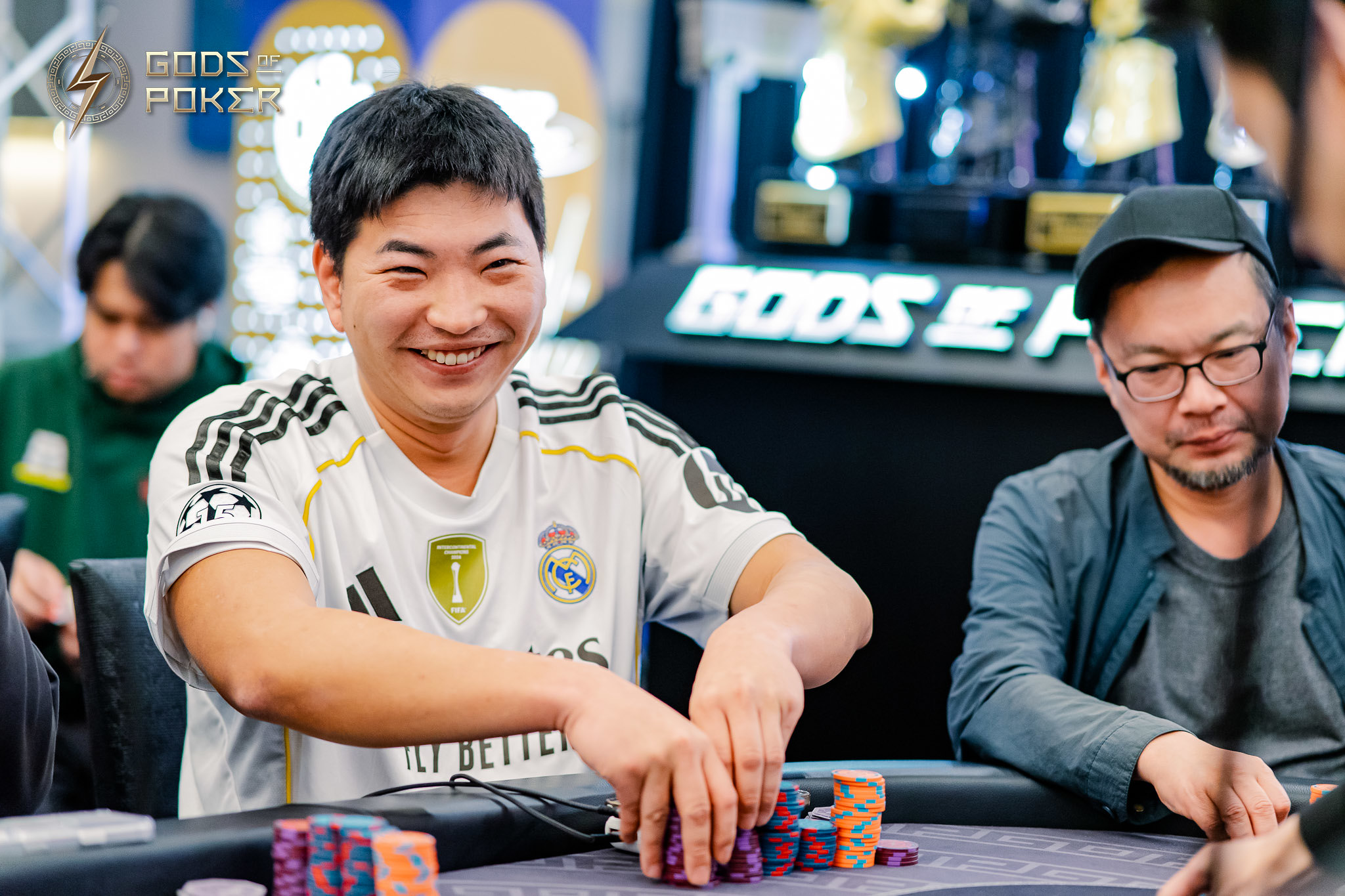 Smiling man in a white jersey pushes poker chips forward at a brightly lit casino table.