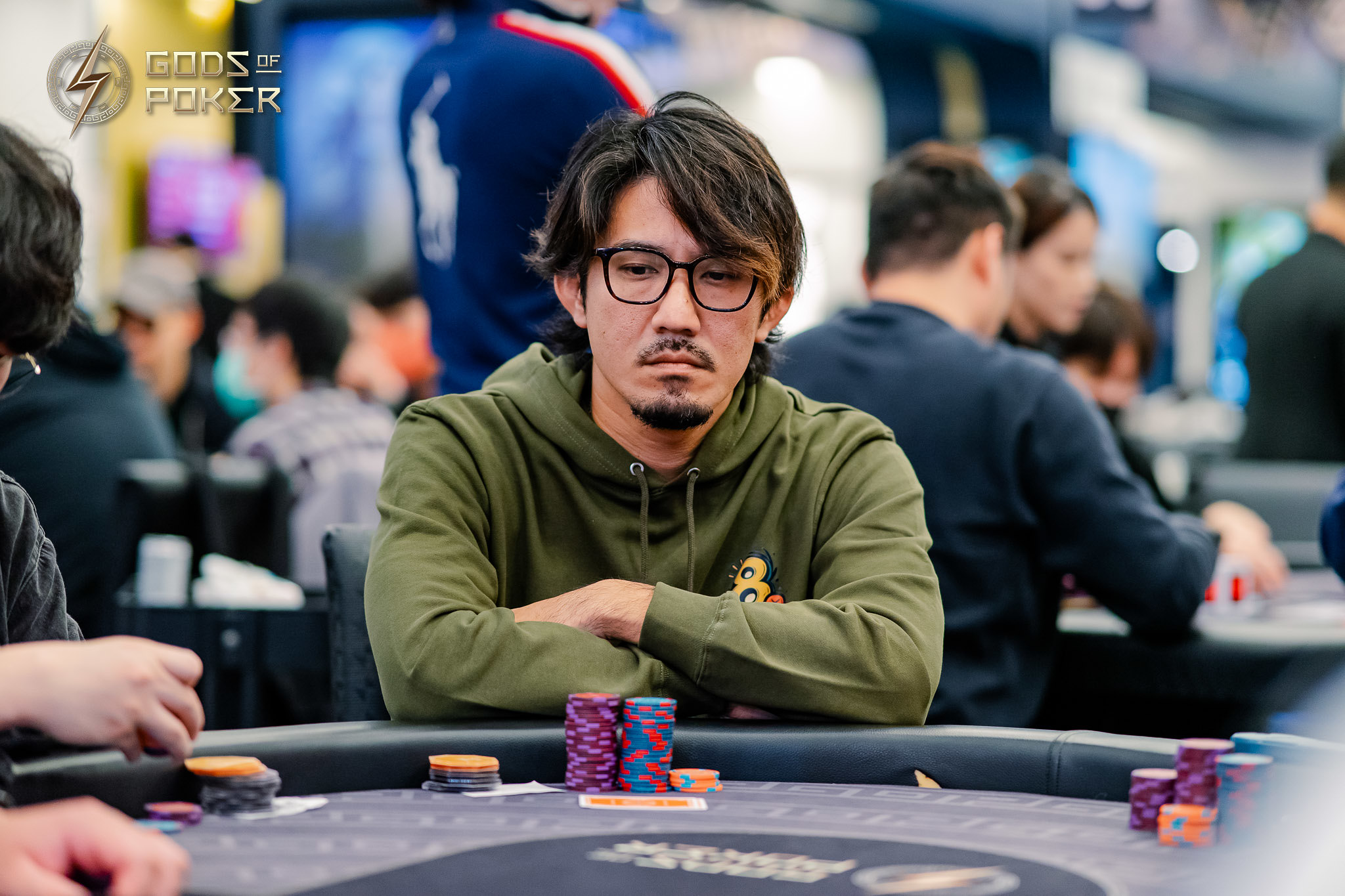 Man with glasses and green hoodie sits intently at a poker table with chips.