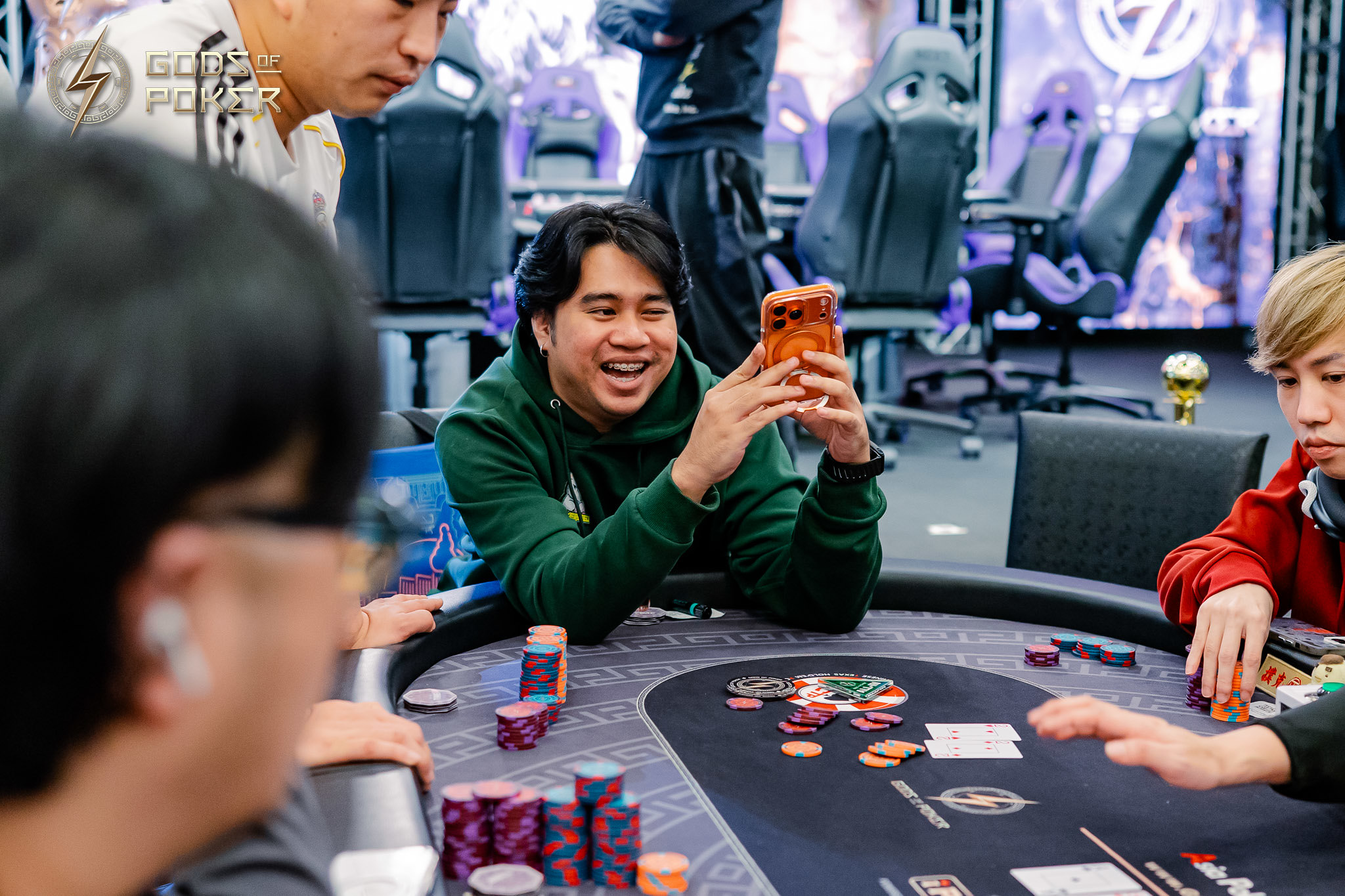 Smiling man in green hoodie checks his phone at a poker table with chips and cards.