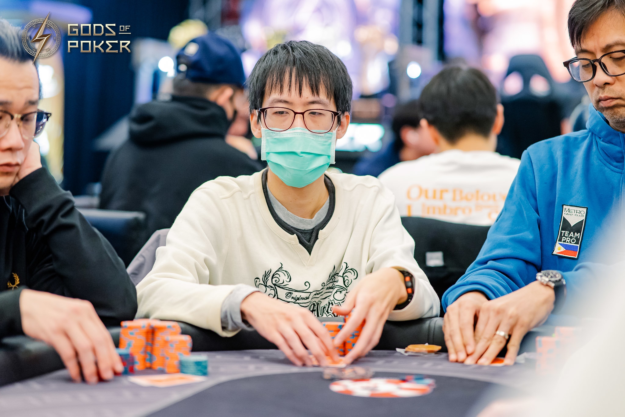 Asian man wearing glasses and a medical mask plays poker, chips on the table.