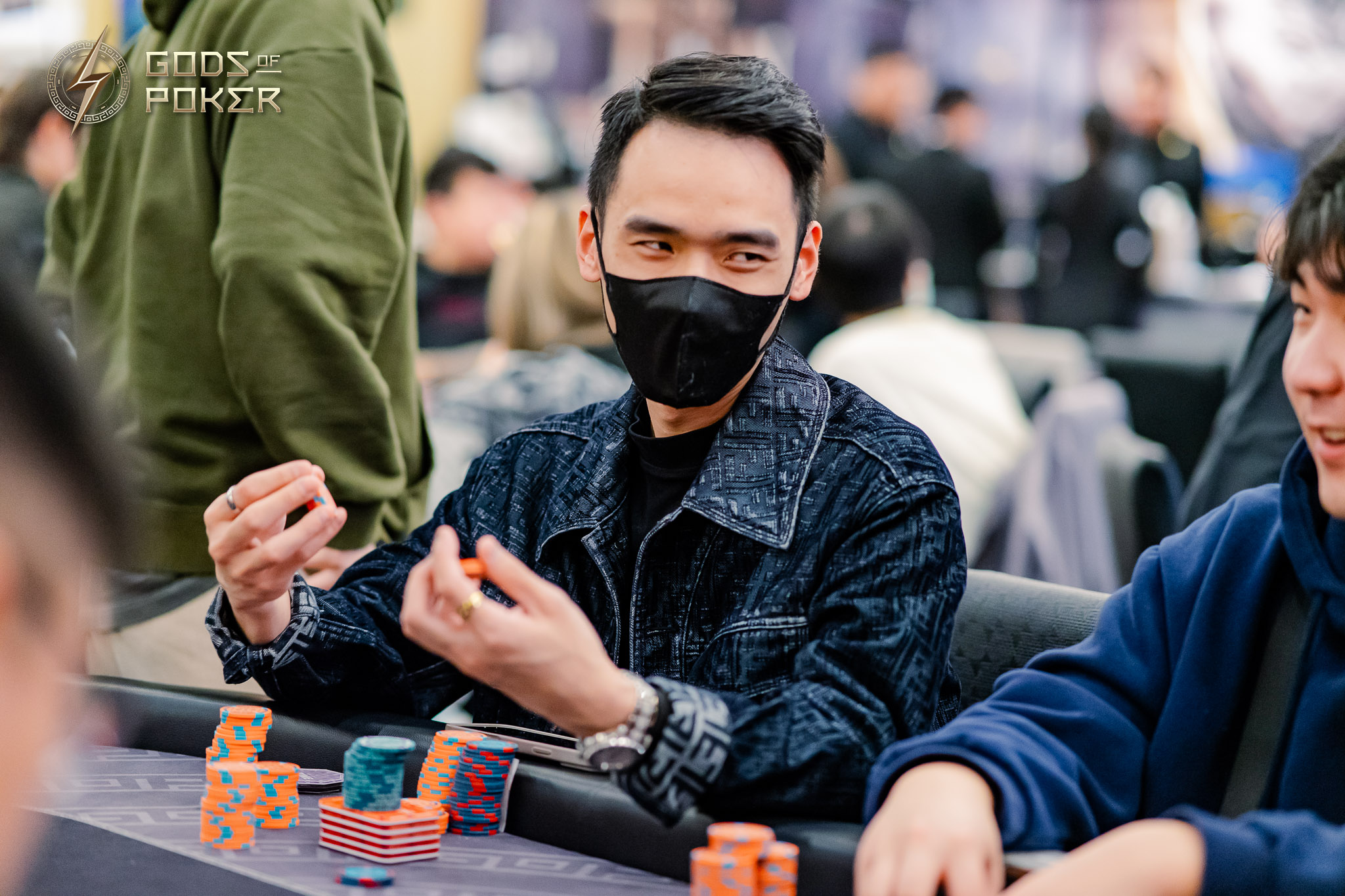Man in black mask and denim jacket gestures at a poker table with chips.