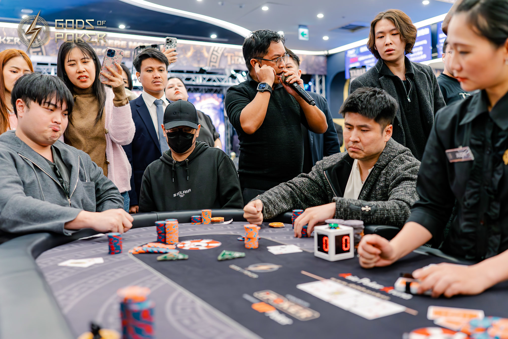 Players and spectators intently watching a poker game at a casino table, with a man speaking.