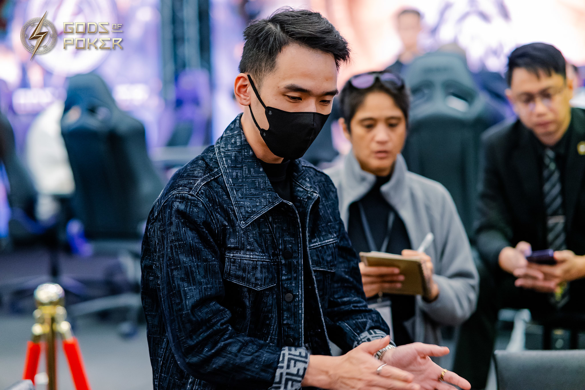 Man in a black face mask and dark denim jacket looking down with hands clasped.