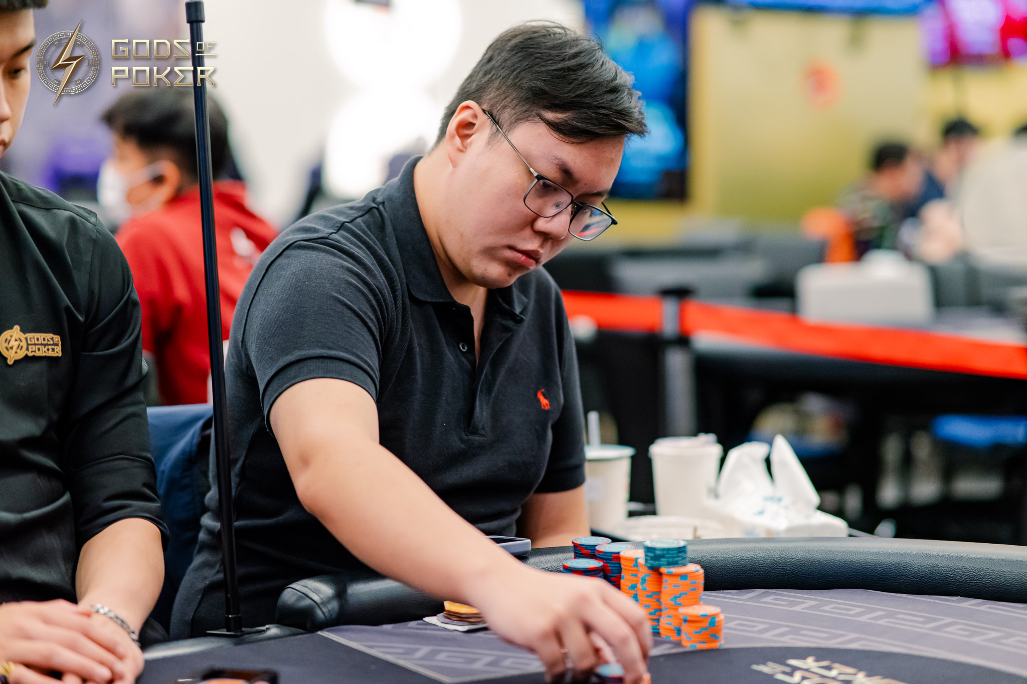 A man in a black polo shirt and glasses plays poker at a casino table.