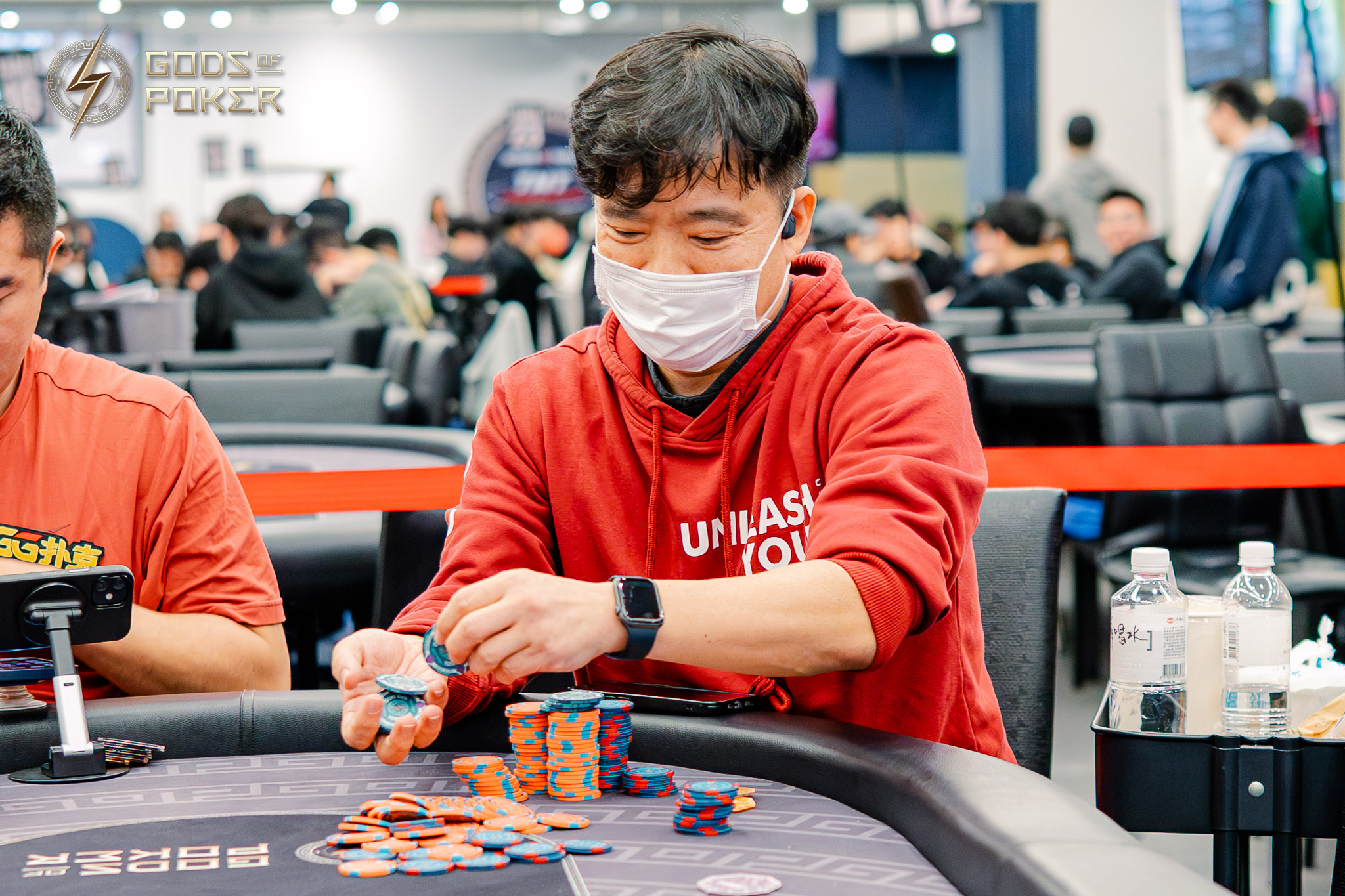 Man in a red hoodie and mask, playing poker and moving chips at a table.
