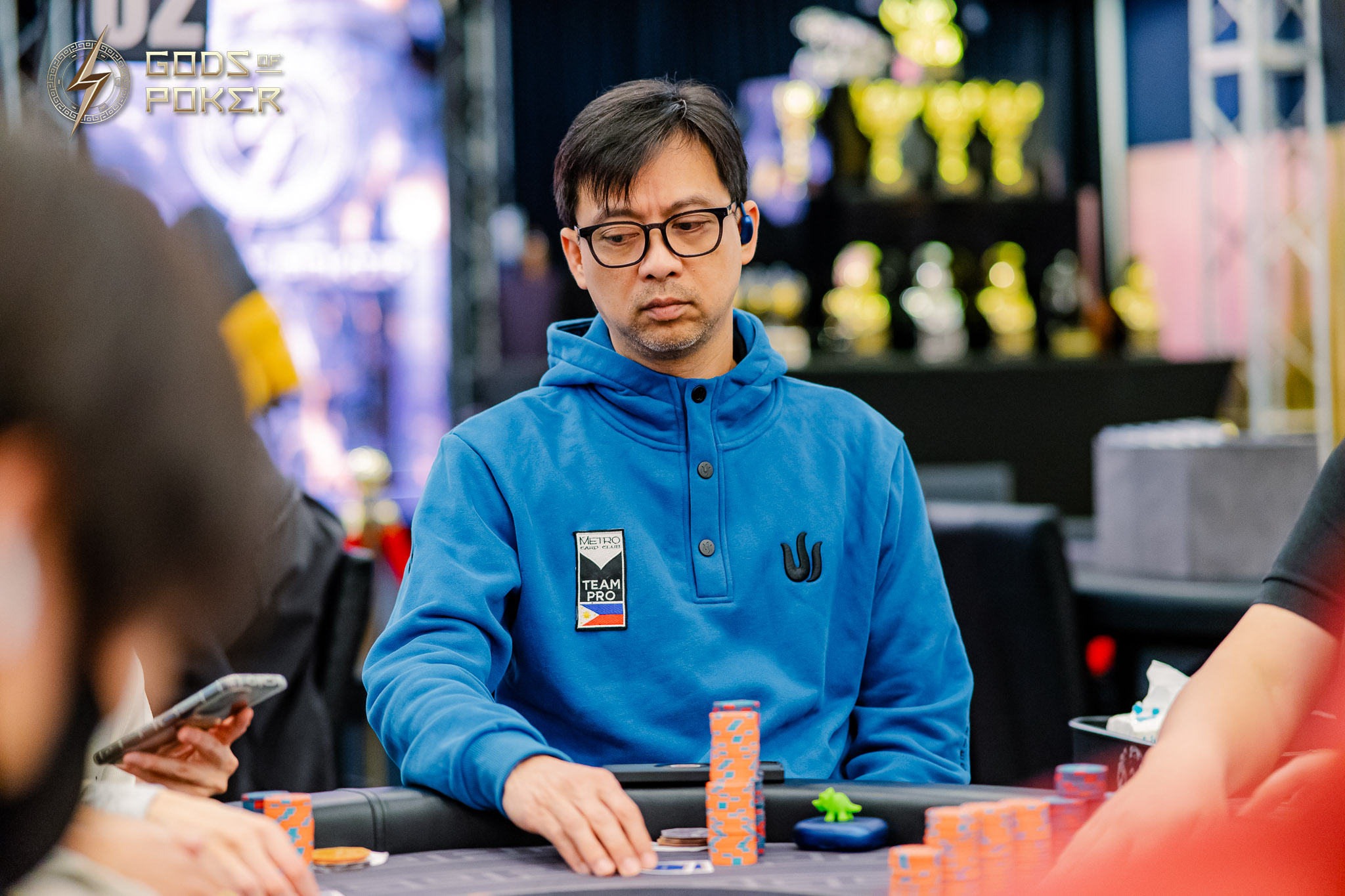 A man in glasses and a blue hoodie seriously contemplates his chips at a poker table.